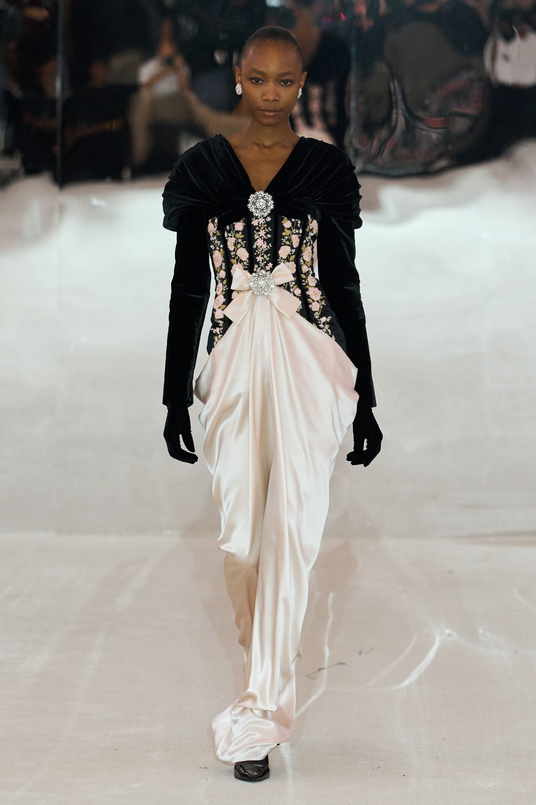 Round crystal cluster earrings. Black velvet bodice with pink floral embroidery and a jewel clasp. Blush satin drape gathered into a bow, falling in heavy folds. Black opera gloves in matte stretch fabric. Black patent square toes peeking from the hem. Crossing a hotel ballroom after midnight, the ice sculpture already sweating.