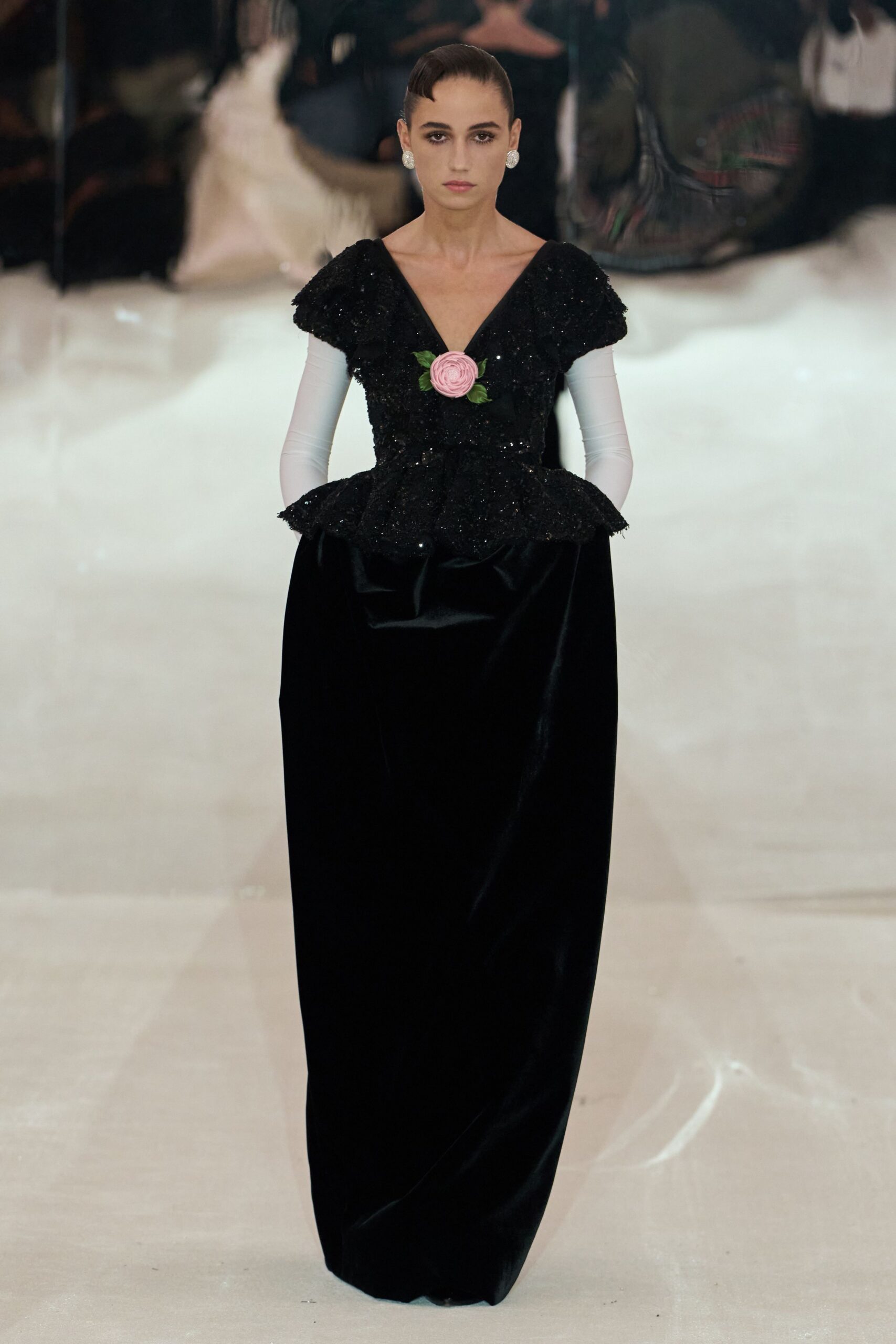 Round crystal cluster studs. Black sequined bodice with cap sleeves and a pink rosette at the plunge. White stretch gloves reaching past the elbow. Floor-length black velvet column, peplum flaring sharply at the hip. Crossing a hotel ballroom after the orchestra has packed away.