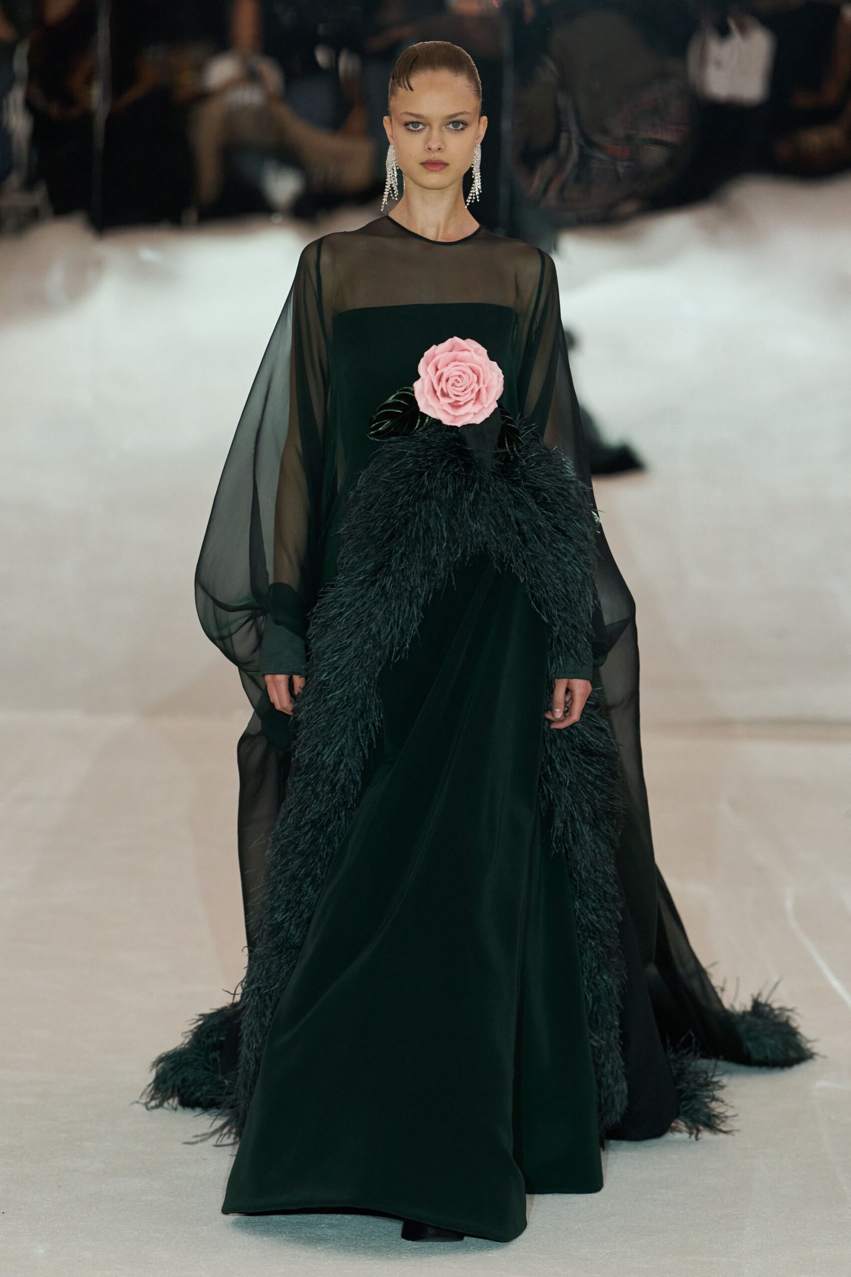 Long crystal fringe earrings brushing the jaw. Black floor-length gown with a sheer yoke and balloon chiffon sleeves. Pale pink rose fixed high at the waist with glossy leaves. Black feather edging climbing the hips and trailing into the hem. At the charity ball after dinner plates are cleared and the quartet starts again.