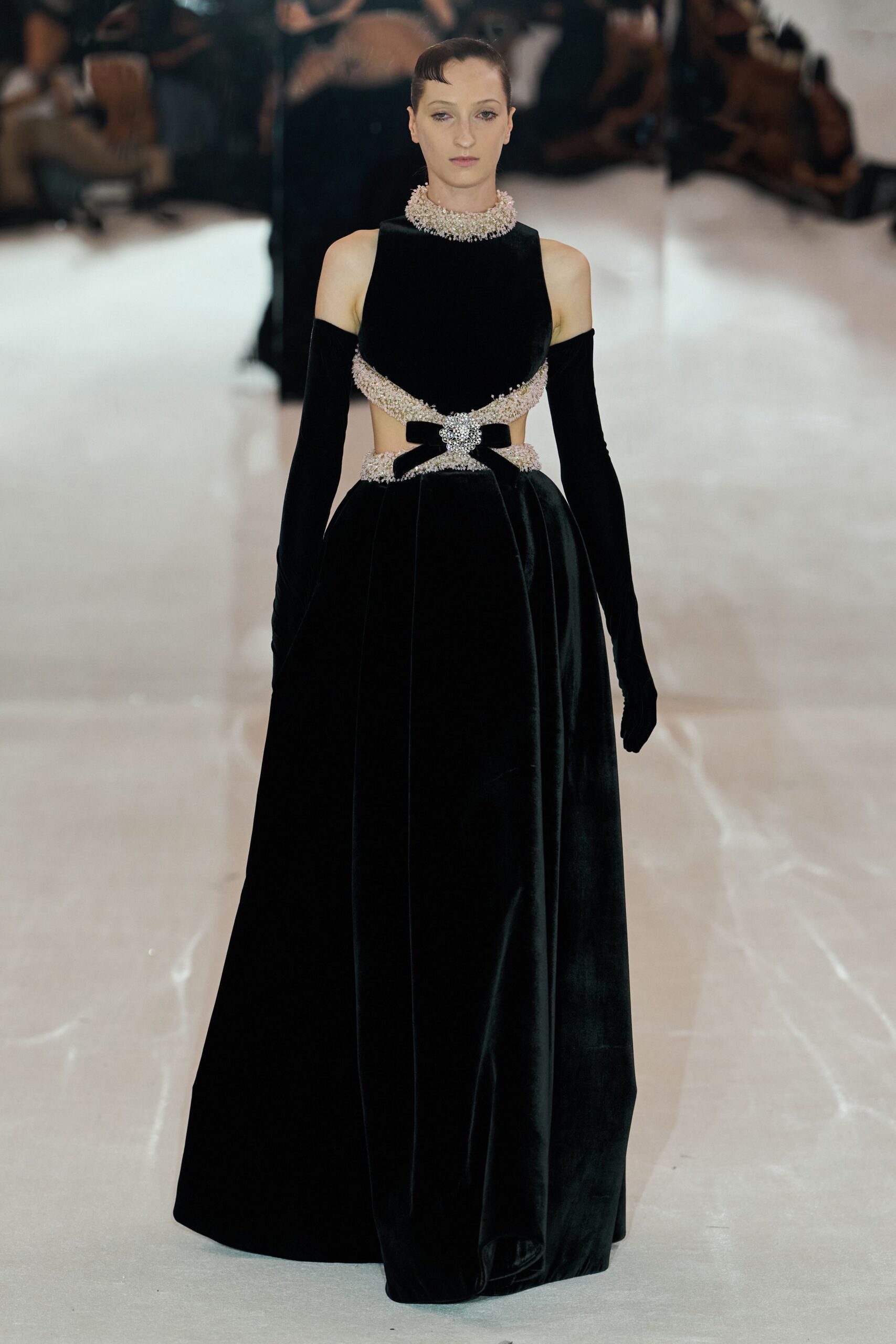 Black velvet gown with a cutout bodice and pooled floor-length skirt. Pale crystal-beaded collar packed tight around the neck. Black velvet opera gloves slipping off the shoulders. Velvet bow at the waist, pinned with a clustered jeweled buckle. For crossing a hotel ballroom after the auction has finally ended.