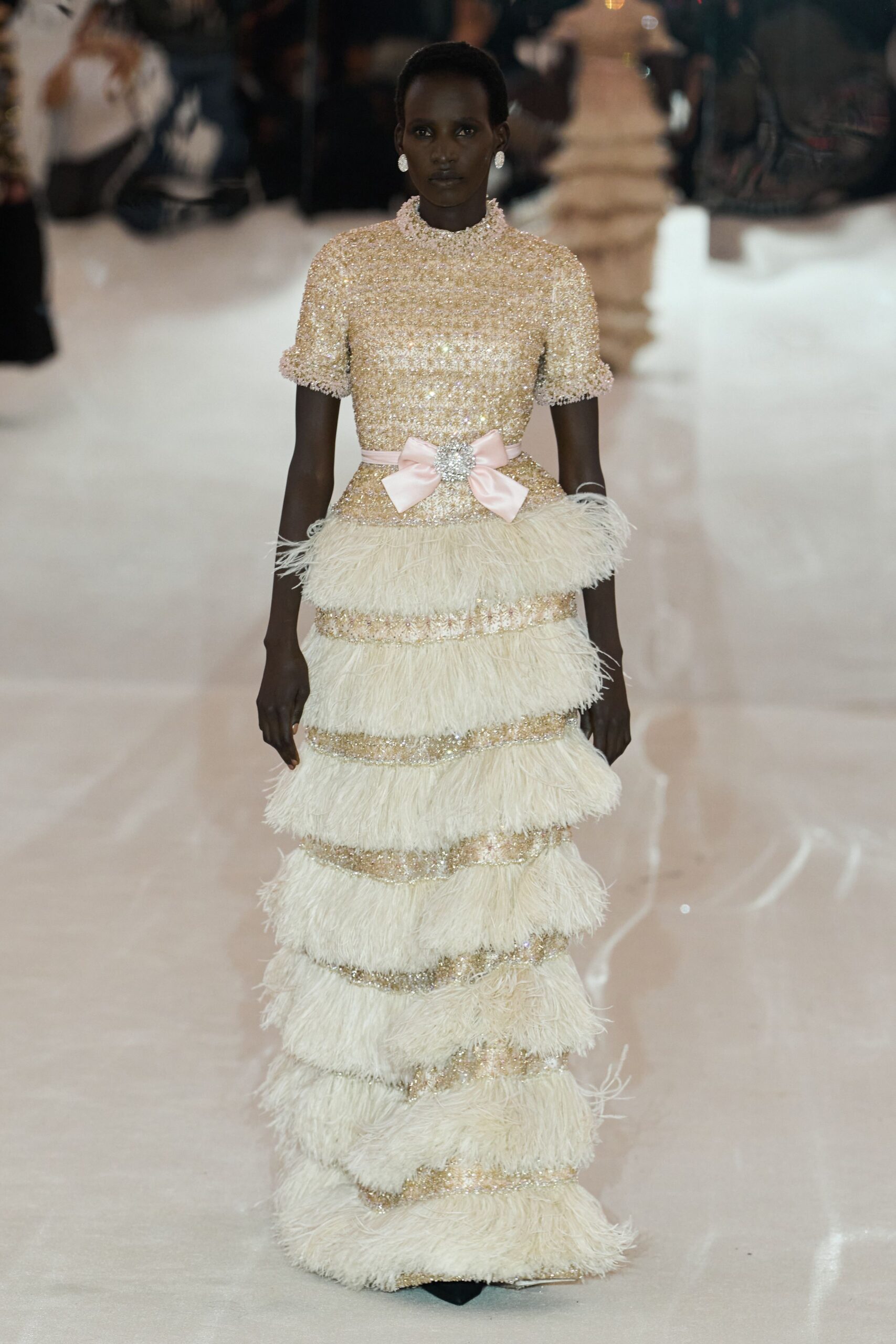 Round crystal clip earrings. Gold beaded column gown with puffed cuffs and ostrich-fringe tiers. Blush satin bow belt centered by a rhinestone cluster. Black pointed toes just breaking the hem. Hotel ballroom carpet rolled back for a wedding that started at noon.