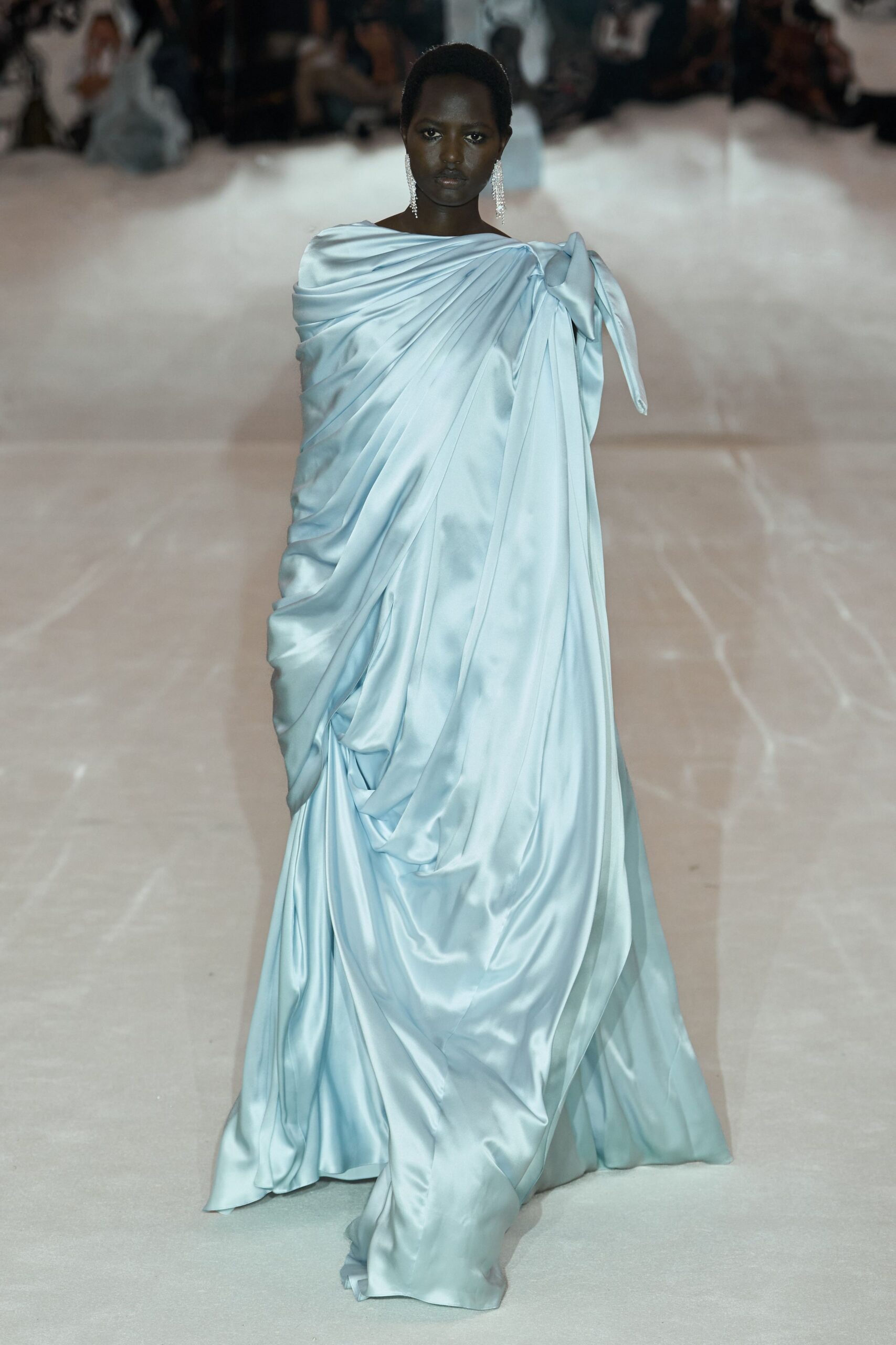 Ice-blue satin gown draped in diagonal folds, one shoulder tied into a long bow. Crystal fringe earrings brushing the jaw. Crossing a hotel ballroom after the wedding cake has been cut.