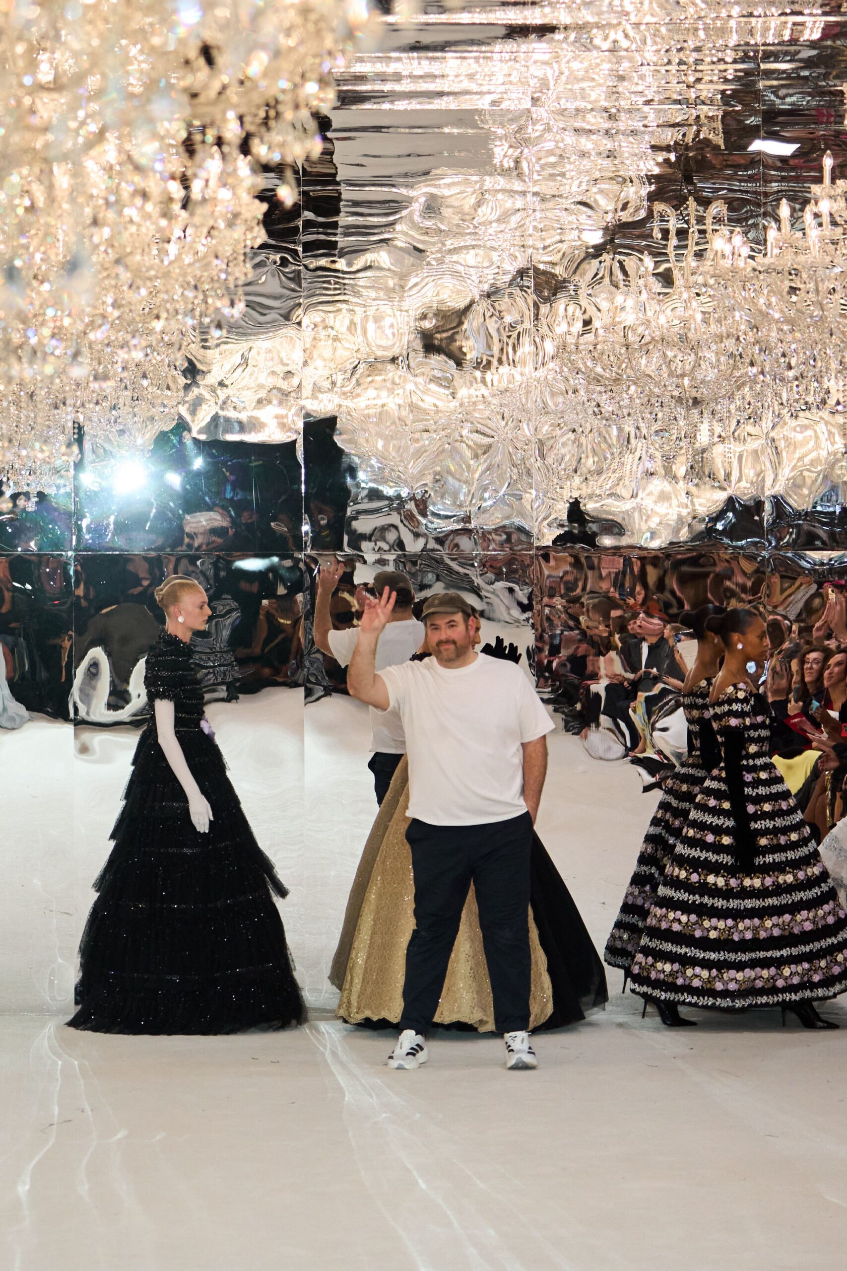 Olive flat cap with a short curved brim. Plain white jersey tee, cut loose through the torso. Dark straight trousers breaking softly above the shoe. White leather sneakers with three black side stripes. Taking a bow under mirrored chandeliers while everyone else is still in gowns.