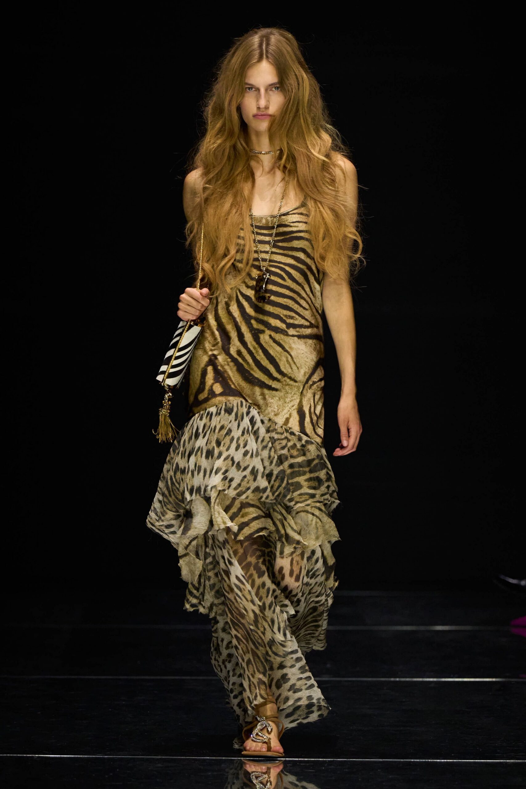 Bias-cut slip dress mixing zebra and leopard, chiffon hem breaking into loose ruffles. Long chain with a dark pendant hanging to the sternum. Zebra shoulder bag with a gold chain strap and tassel. Flat metallic sandals with a coiled snake across the toes. Walking back from the beach bar after dark, sand still stuck to one heel.