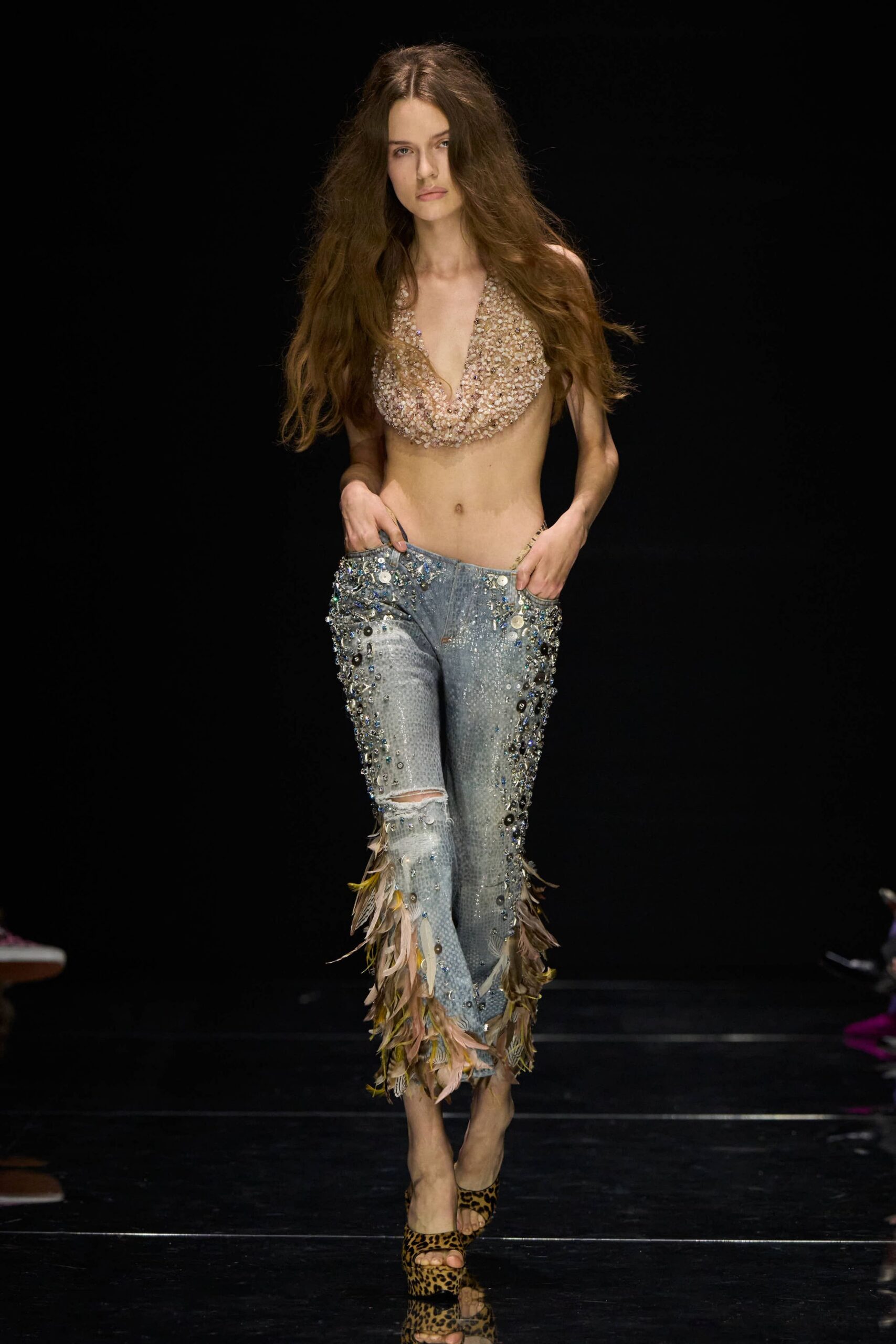 Nude halter bib densely crusted with shells and pale beads. Bleached denim jeans, sequined through the thigh, one knee slit, feathered hems. Leopard pony-hair peep-toe mules on a high platform. Walking off a beach boardwalk after buying trinkets you should not have.