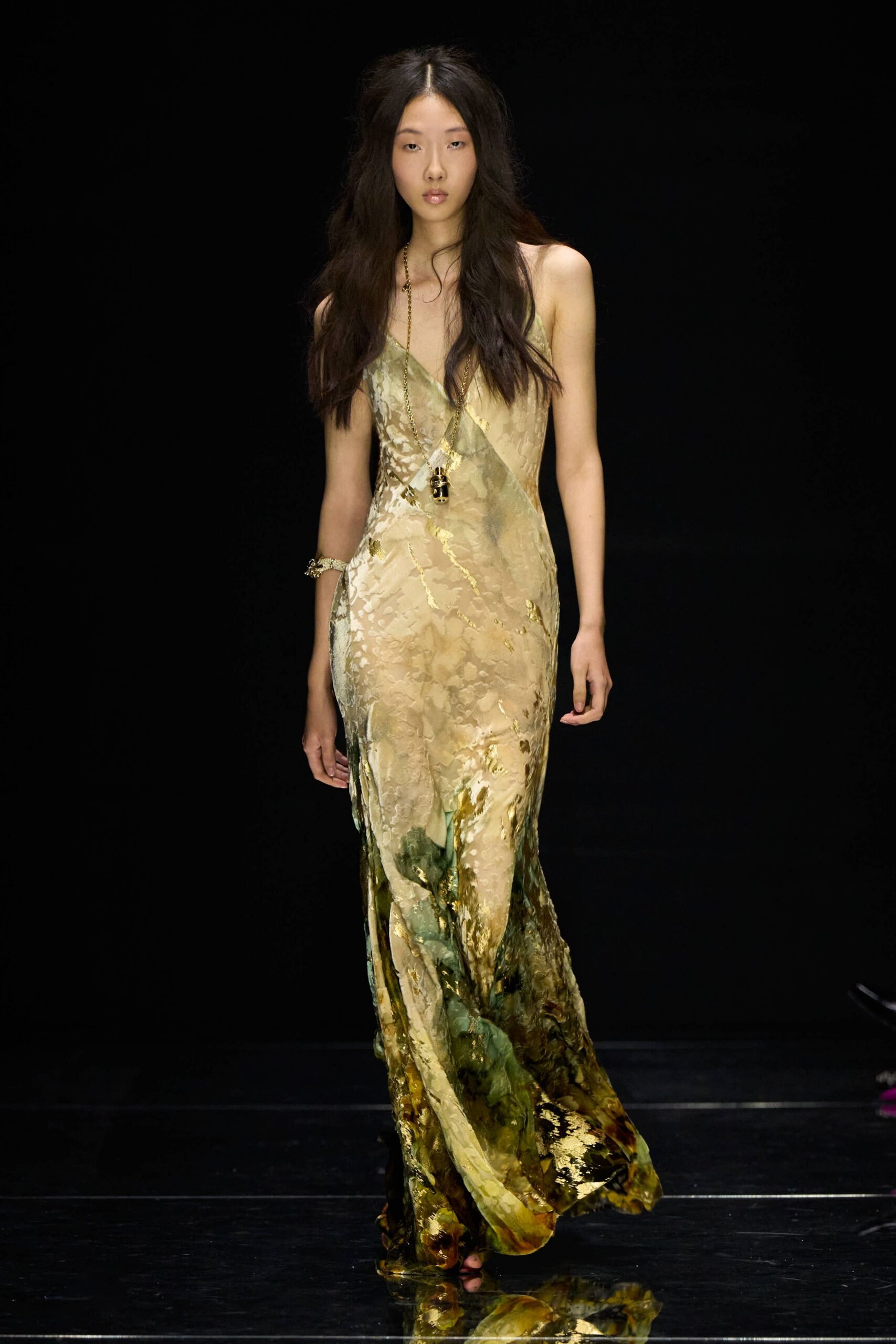 Bias-cut slip dress in mottled celadon and sand devore, gold foil catching the hem. Long chain necklace with a beetle-shaped pendant hanging at the sternum. Clustered gold bracelet at one wrist. Greenhouse aisle after rain, palms dripping onto the concrete path.