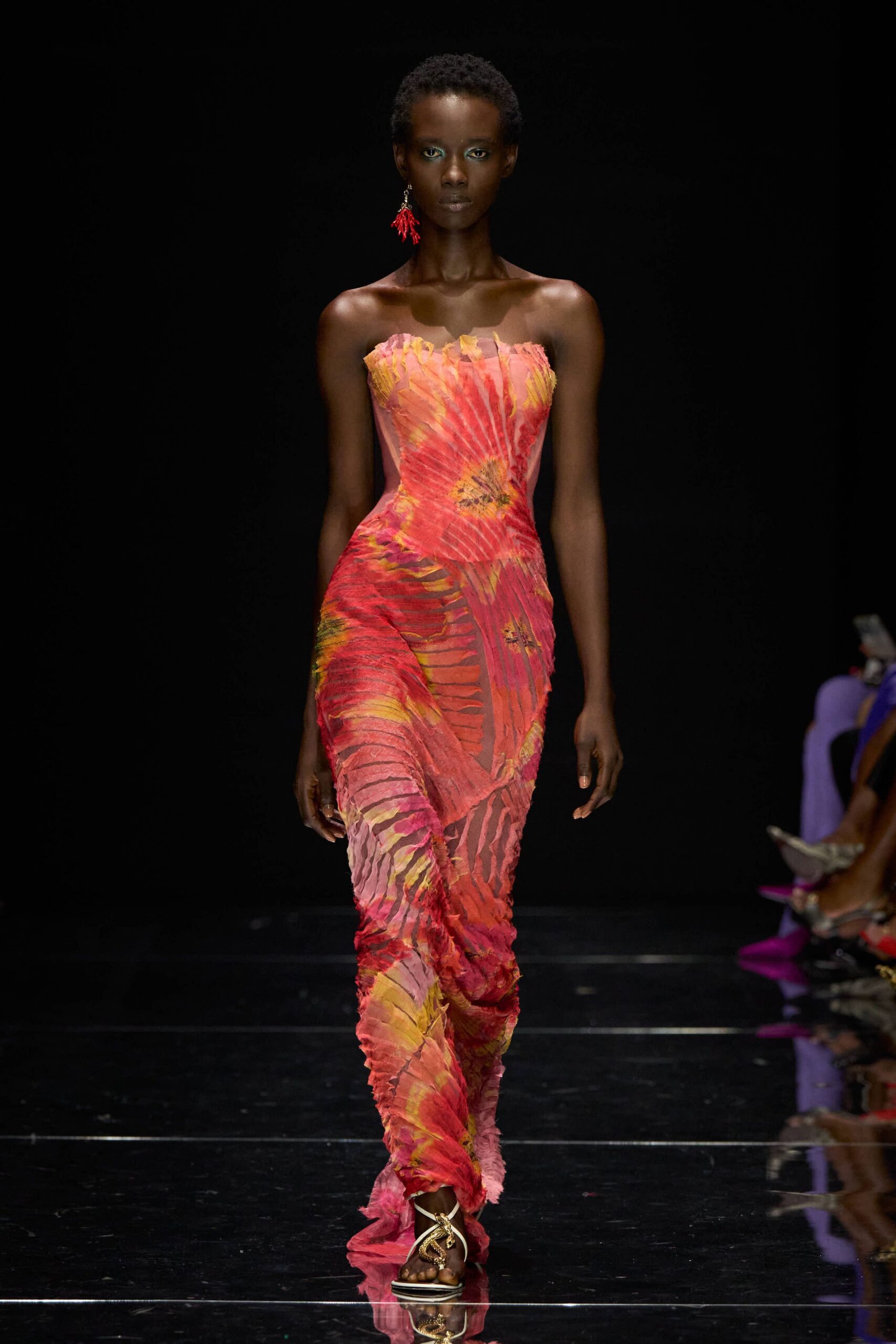 Single coral-branch earring in lacquered red. Strapless column gown in pleated sheer mesh, hibiscus print warped over corseted hips. Gold thong sandals with a coiled serpent across the toes. Walking the length of a hotel pool after the sun has gone but the tiles still hold heat.
