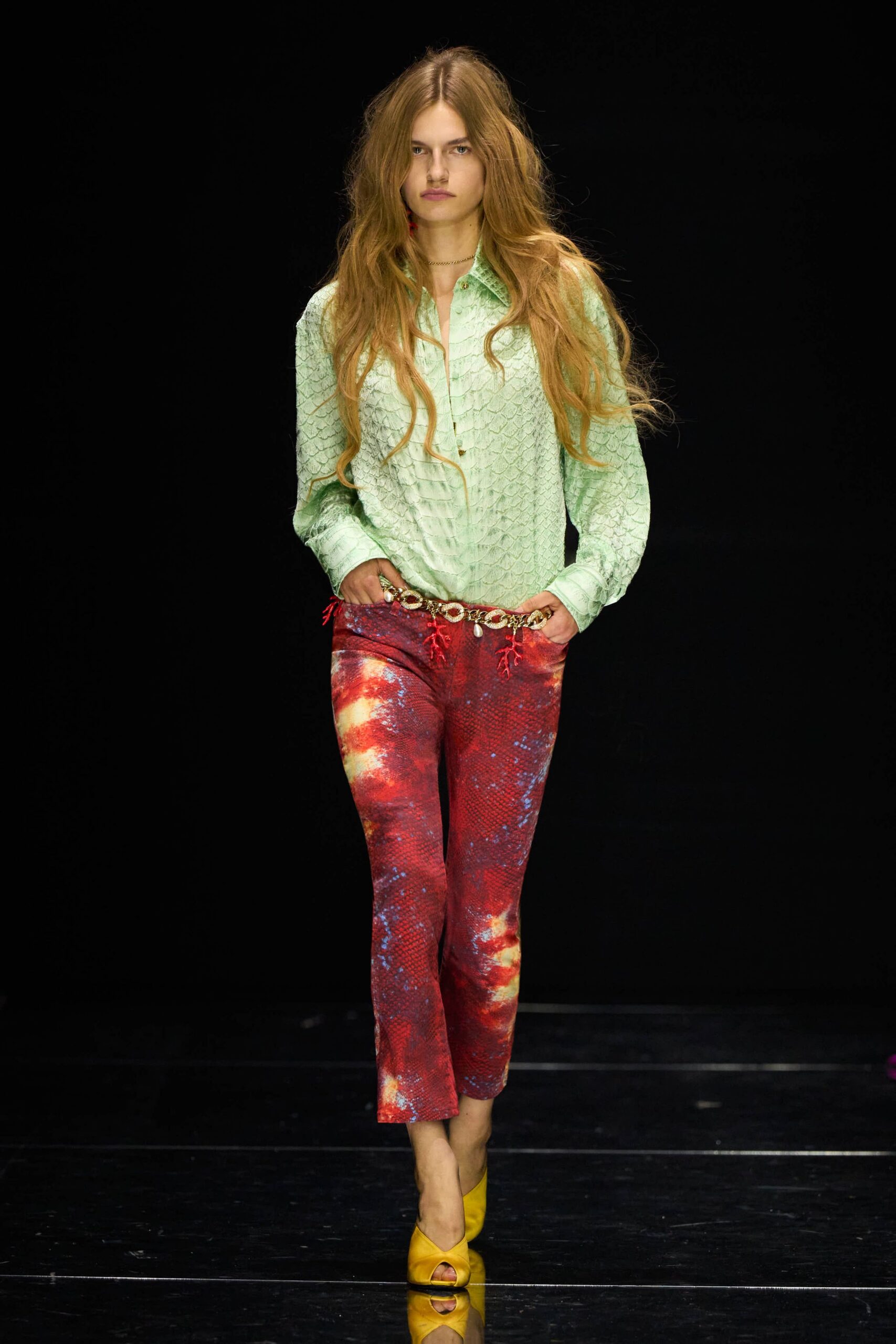 Mint python-textured shirt, collar spread open above the third button. Red snakeskin-print cropped trousers, mottled with cloudy yellow bursts. Gold chain belt hung with coral-red tassels and a pearl drop. Mustard satin peep-toe mules. Walking the seafront after sunset with salt still drying on your wrists.