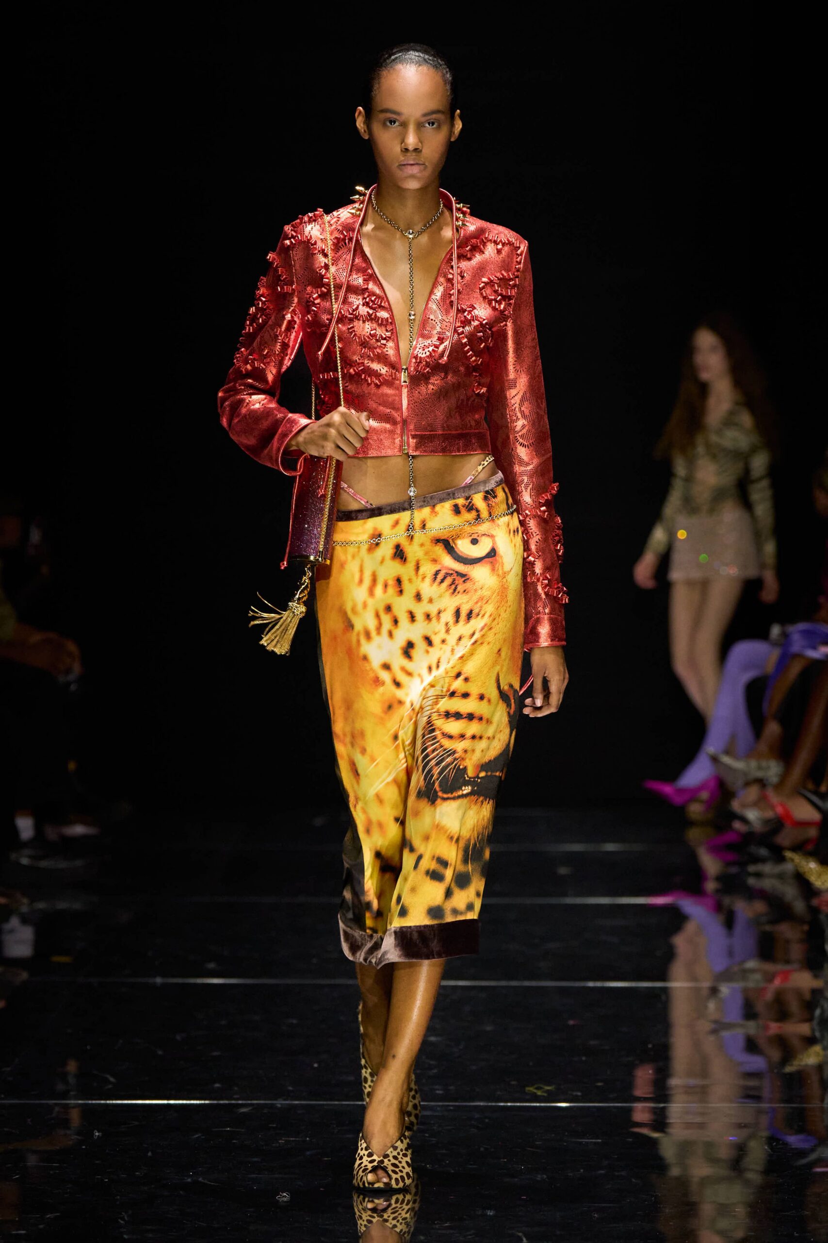 Cropped red satin jacket, laser-cut and tufted into shaggy rosettes. Silver lariat chain dropping straight into the deep zip V. Slip skirt printed with a giant leopard face, velvet hem band. Glitter tube bag with a long gold tassel. Leopard pony-hair peep-toe mules. For arriving at a casino bar before anyone has started losing.