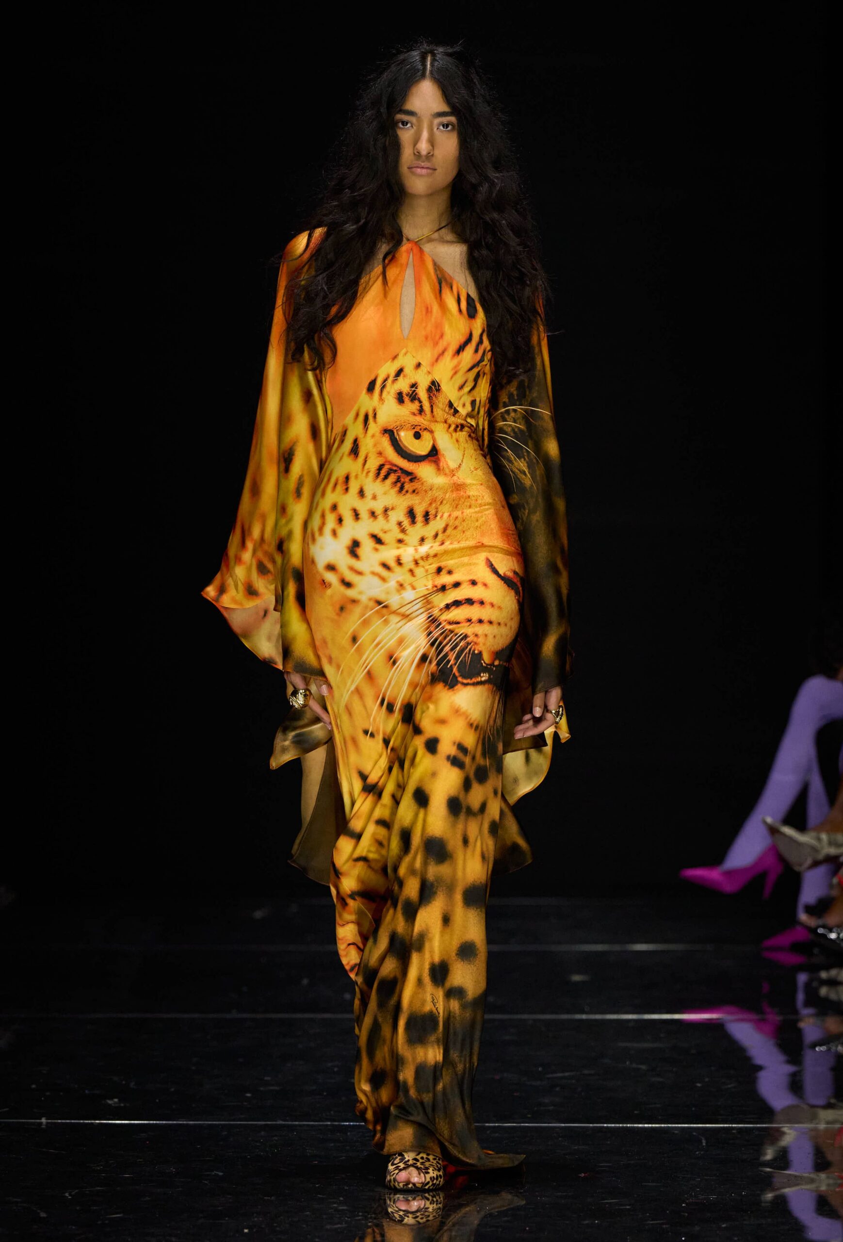 Fluid satin gown printed with a blown-up leopard face, sleeves hanging past the hands. Leopard peep-toe sandals under the pooling hem. Chunky gold rings on both hands. Crossing a casino lobby at sunrise with yesterday's makeup still on.