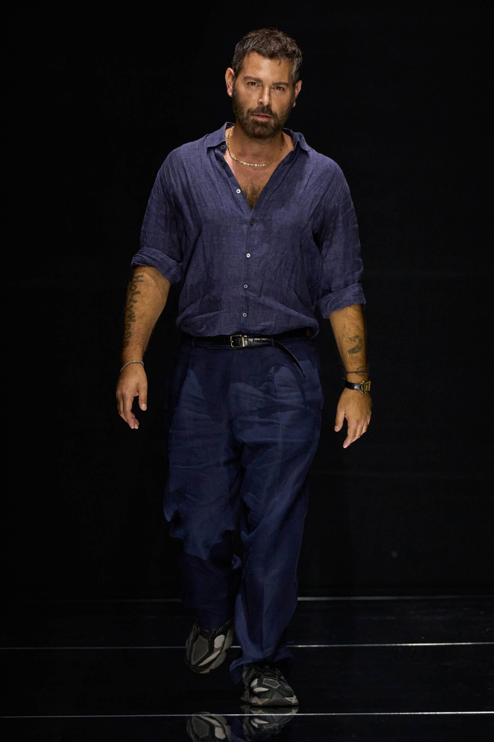 Washed indigo linen shirt, open throat and sleeves rolled bluntly. Loose midnight trousers with deep front pleats and a belt tail hanging. Charcoal panelled trainers with thick grey pods at the sole. Walking to the harbor restaurant after the heat finally broke.