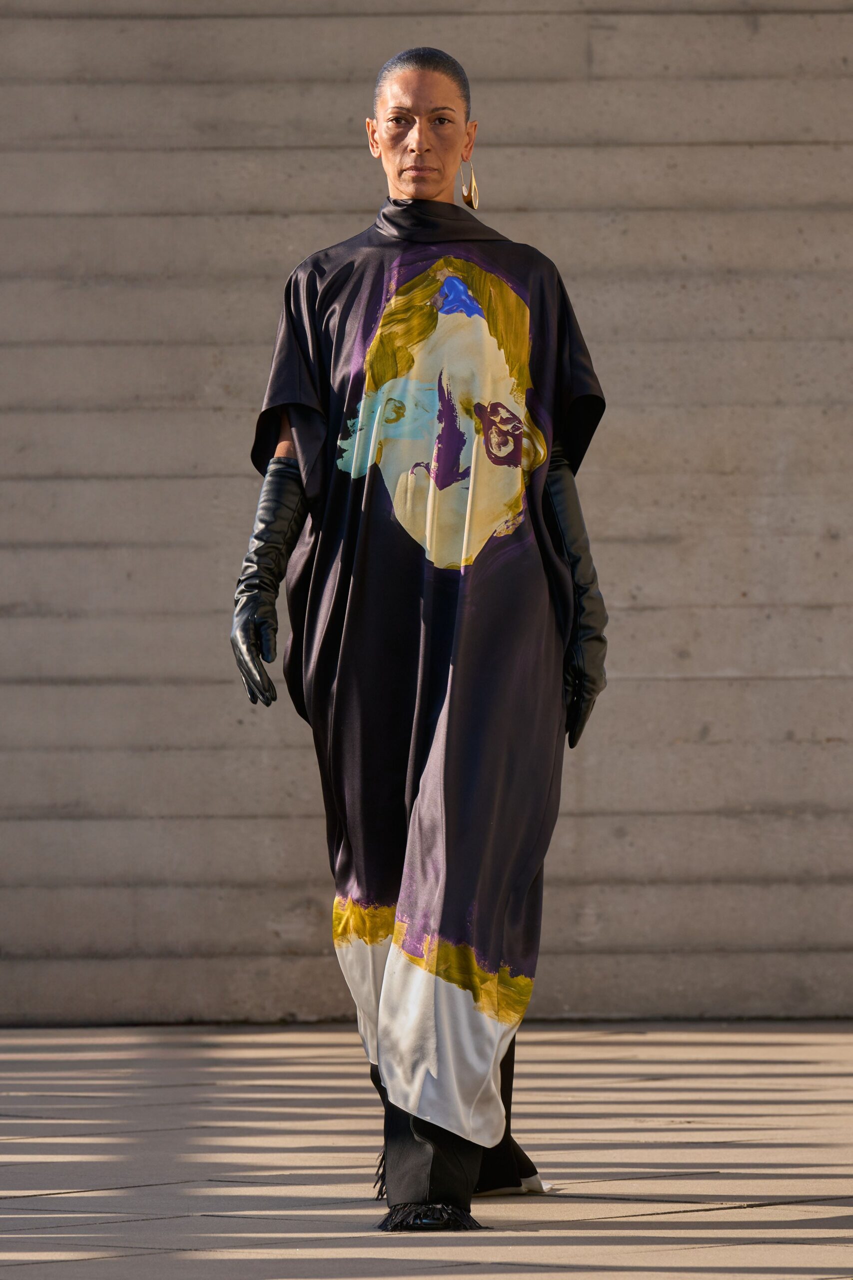Inky satin caftan, mock neck and giant brushstroke face print. Black leather opera gloves, softly wrinkled through the forearm. Single gold teardrop hoop. Black boots with a little fringe brushing the floor. Crossing a sunstruck courtyard before the museum doors unlock.