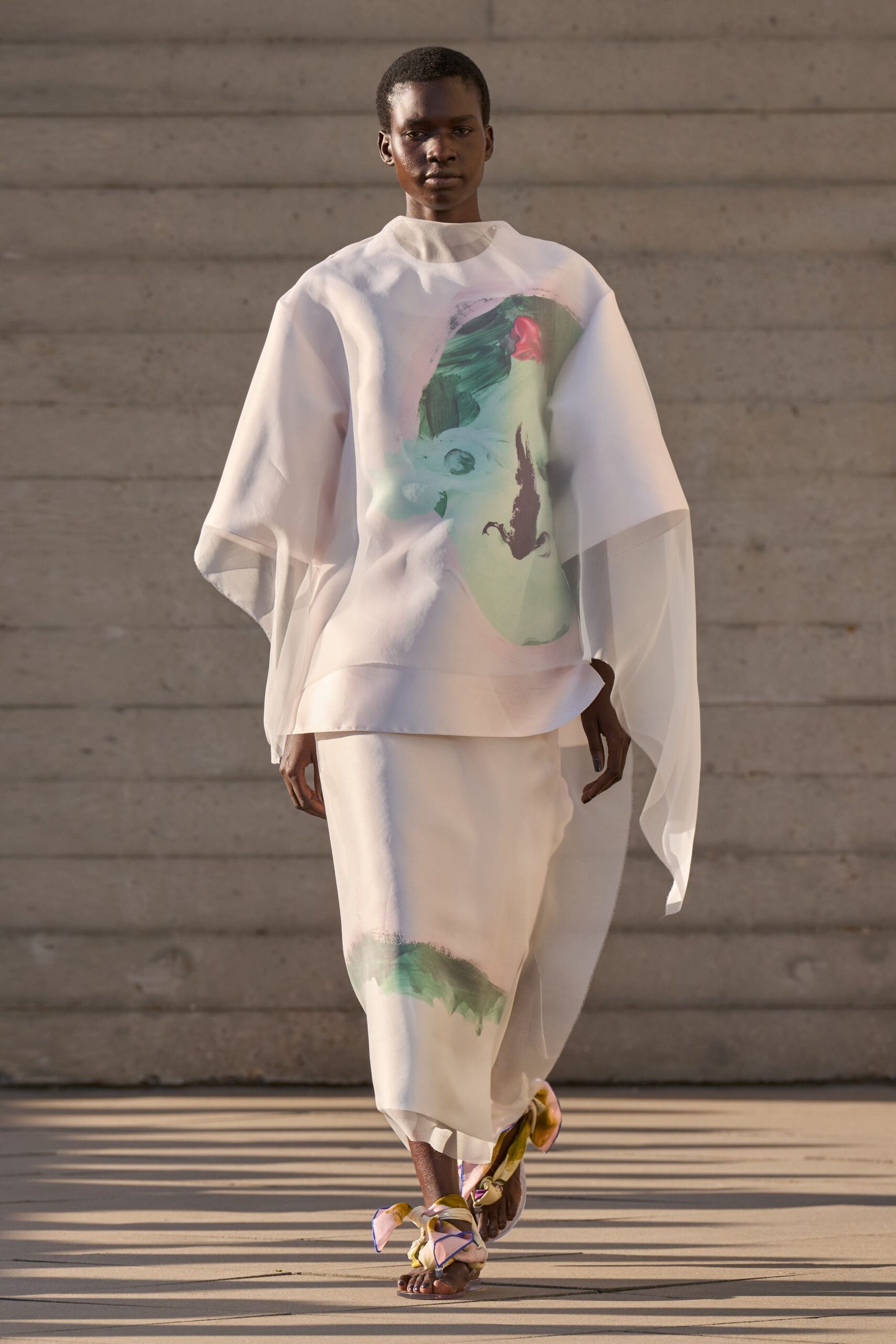 Sheer white organza tunic with a high neck and brushstroke print. Matching column skirt, translucent hem floating past the calf. Flat sandals tied with oversized pastel satin knots. Crossing a museum courtyard just before the heat lifts off the stone.