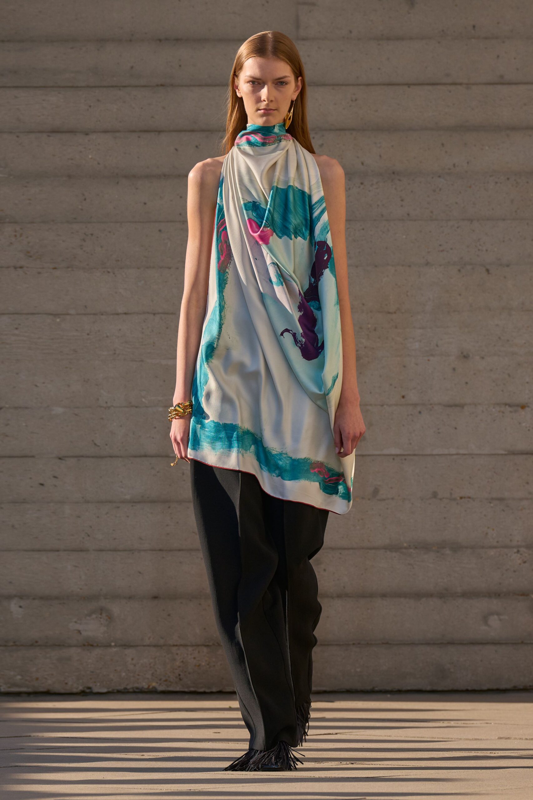 Ivory satin halter tunic, brushstroke print pooling into an uneven hem. Black crepe trousers falling long over shoes with fringe at the cuff. Stacked gold bangles. Single elongated gold drop. Sun on poured concrete, crossing the museum plaza before it closes.