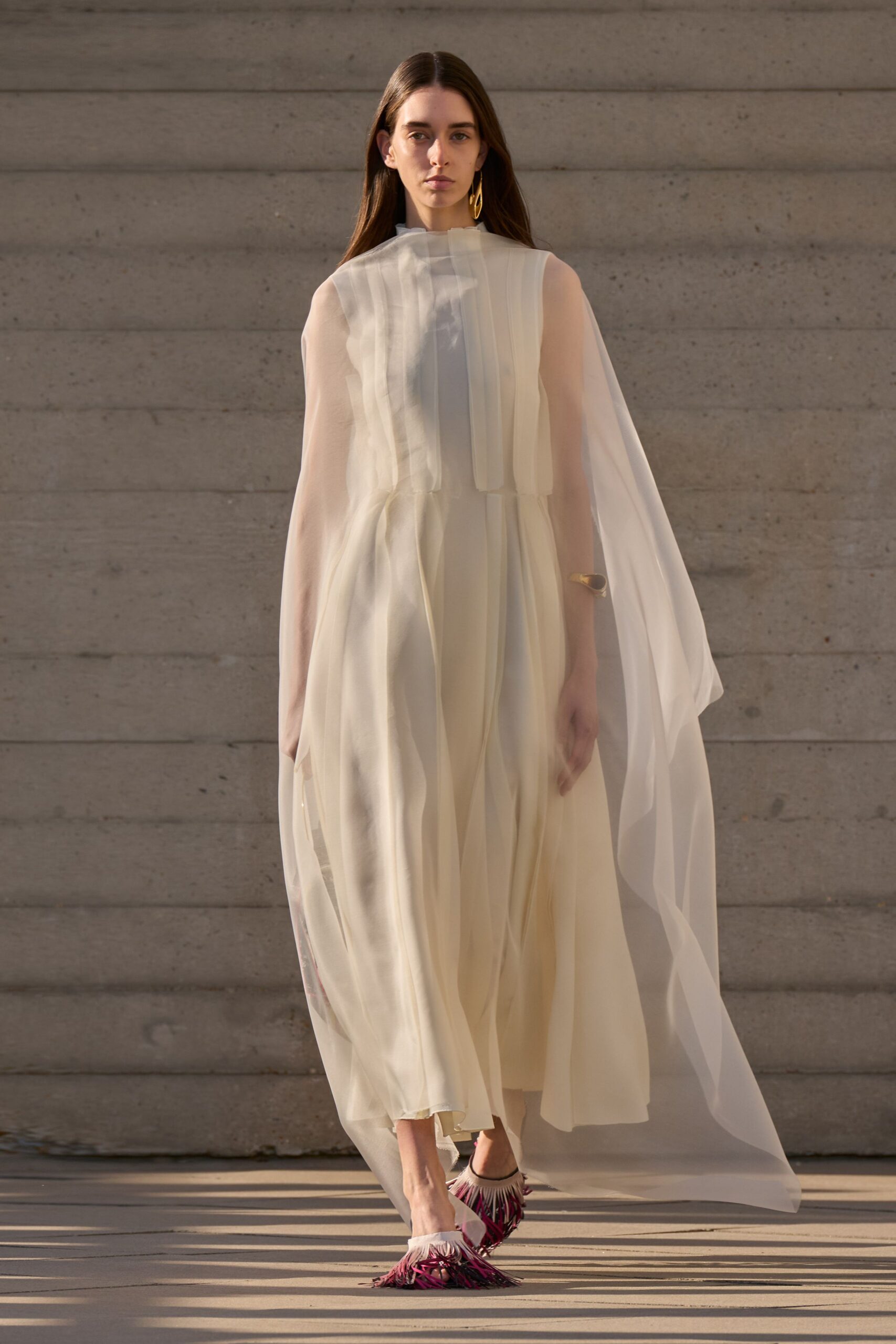 Ivory chiffon cape dress with sheer floor-sweeping panels and narrow pintucks. Single gold drop earring. Brushed gold cuff, worn low on the wrist. Fringed sandals in magenta and black, white ruffle at the vamp. Crossing a museum courtyard just before the heat lifts.