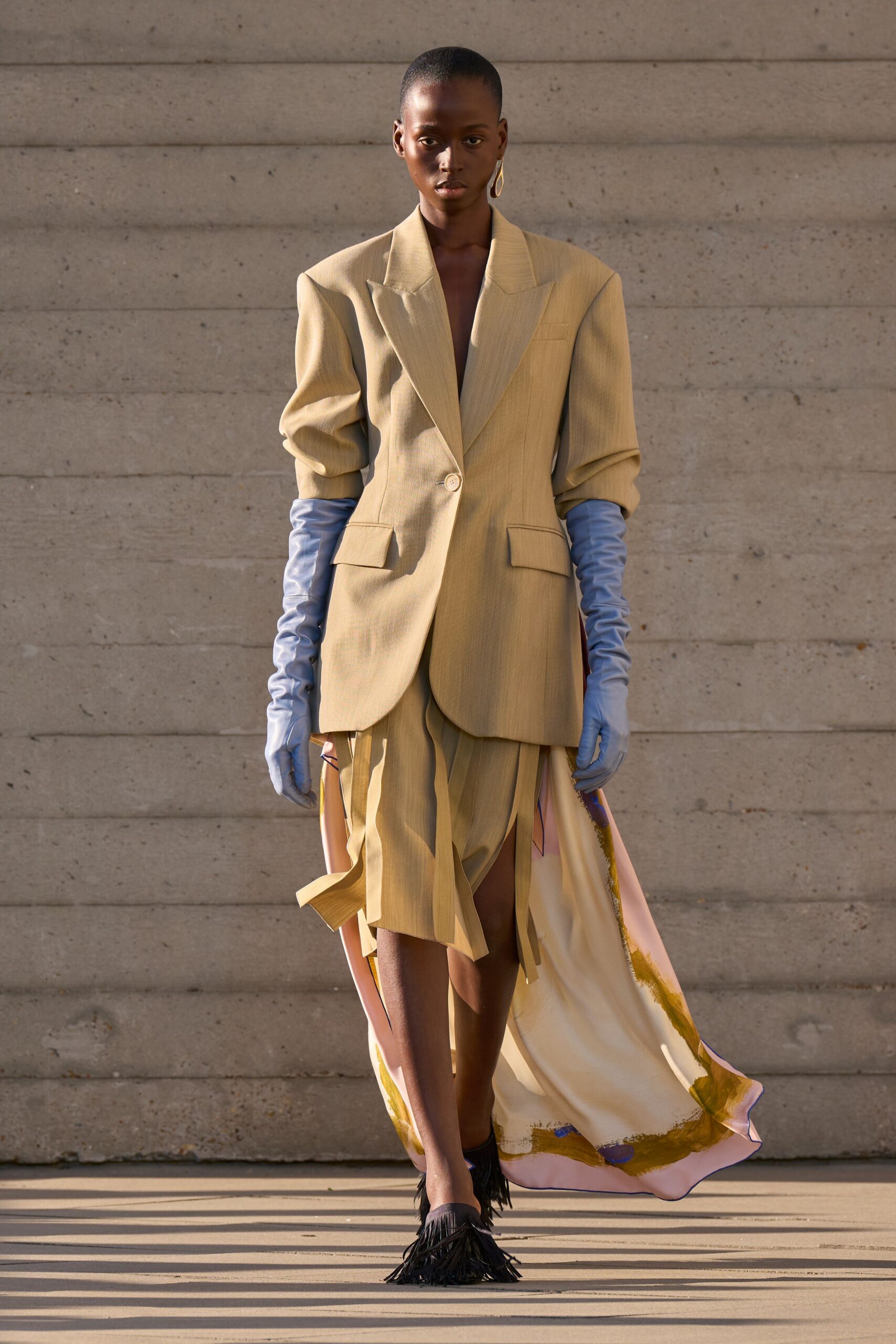 Sand suiting blazer with peaked shoulders and one pale horn button. Powder-blue satin gloves ruched to the elbows. Asymmetric sand skirt split into hanging strips over a painted silk train. Black flats with long fringe skimming the pavement. Single oval drop earring. Walking across a museum courtyard just before the doors open.