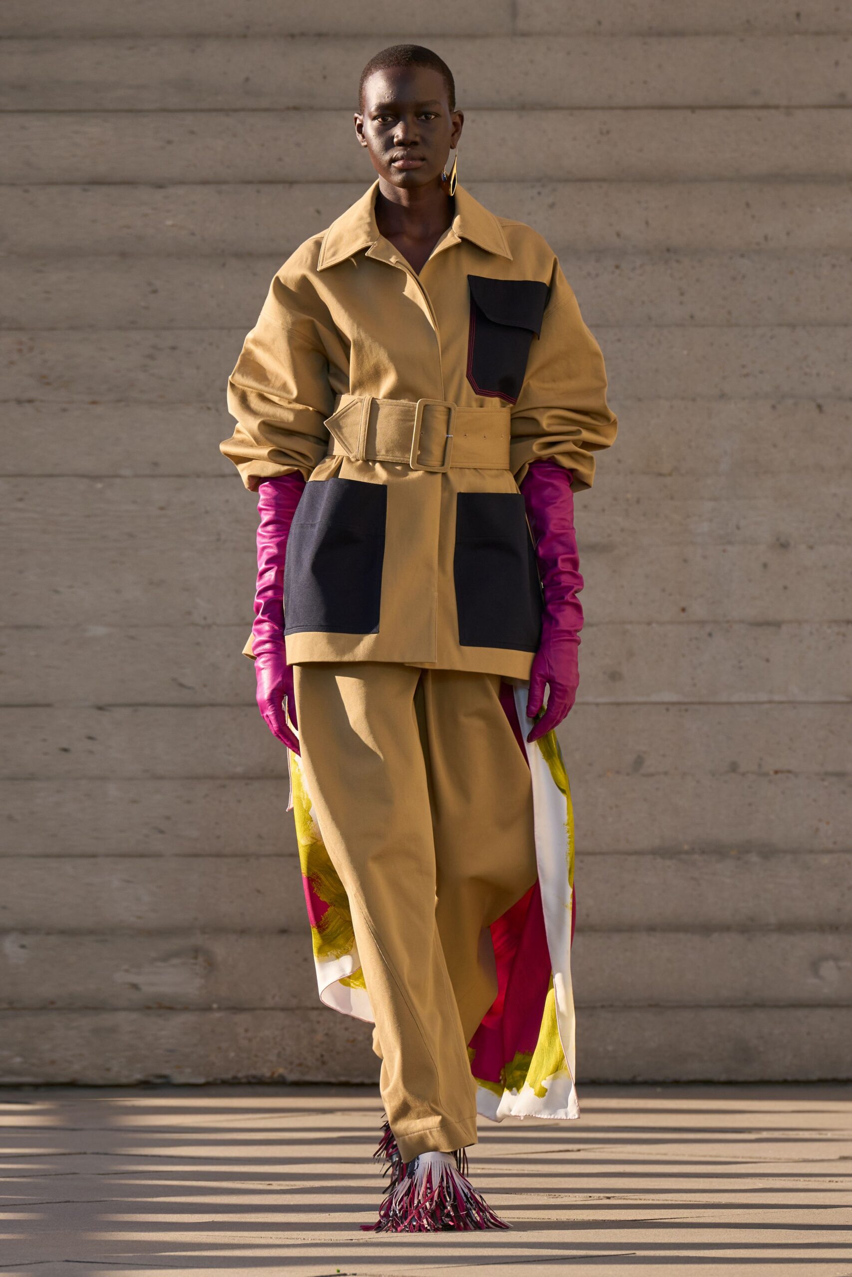 Single gold hoop with a cobalt drop. Khaki cotton coat, huge belt and black patch pockets. Fuchsia satin opera gloves. Wide khaki trousers over a trailing floral panel. Pointed shoes finished in swinging magenta fringe. Crossing a museum courtyard before the fountains switch on.