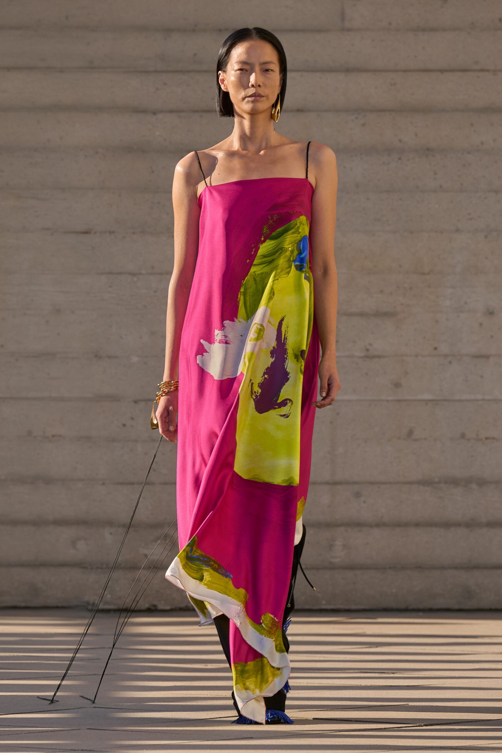 Fuchsia satin slip dress with thin black straps and chartreuse brushstrokes. Stacked gold chain bracelets at one wrist. Single long gold drop earring. Black pointed shoes with cobalt fringe peeking under the hem. Walking across a sculpture court while the sun drops hard against concrete.
