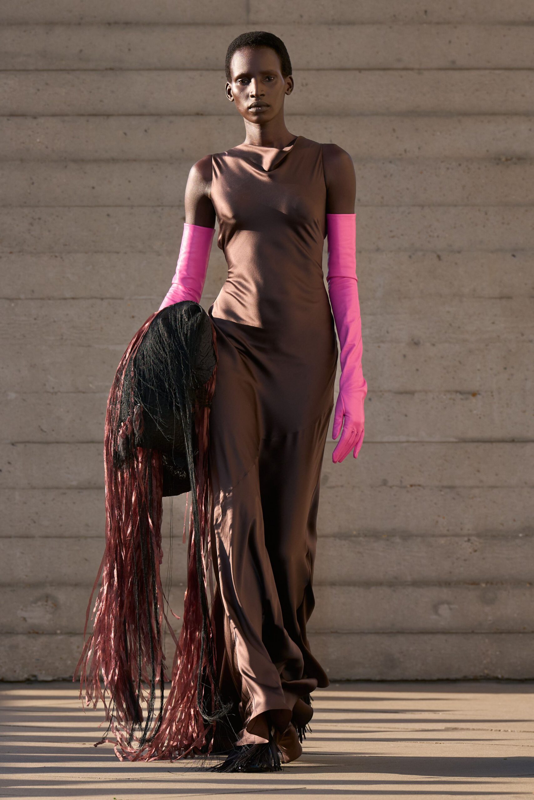 Bronze satin column gown with a soft cowl and twisted side drape. Hot-pink gloves, smooth to the upper arm. Black and oxblood shredded fringe trailing nearly to the ground. Black feathered toes peeking from the hem. Crossing a museum courtyard just before the doors open.