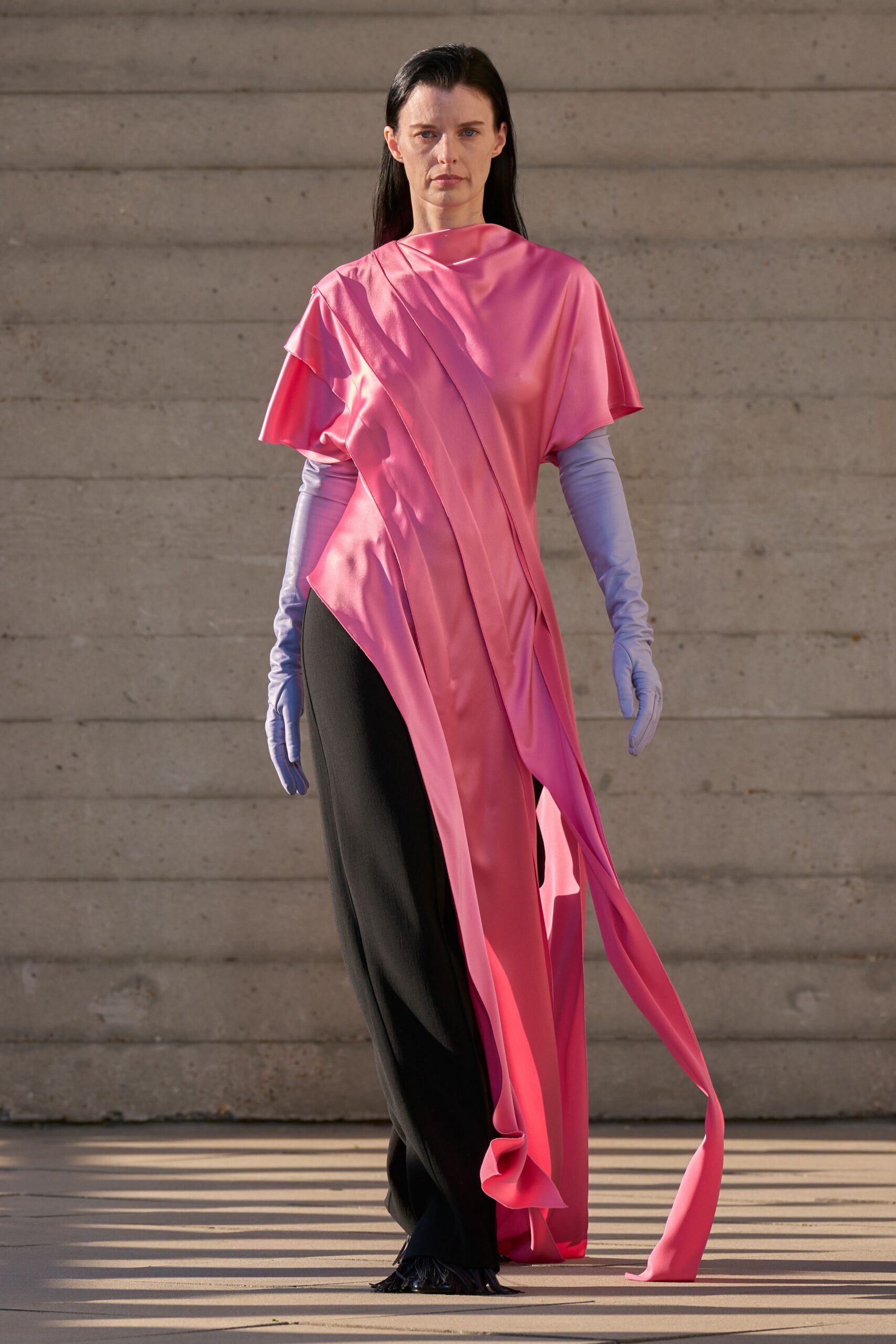 Bubblegum satin tunic draped on the bias, one panel trailing to the ground. Periwinkle opera gloves in a taut stretch finish. Black column skirt falling straight under the split pink drape. Black shoes with a fringe of fine strips at the toe. Crossing a civic plaza before the speeches start, ribbon ends catching dust.