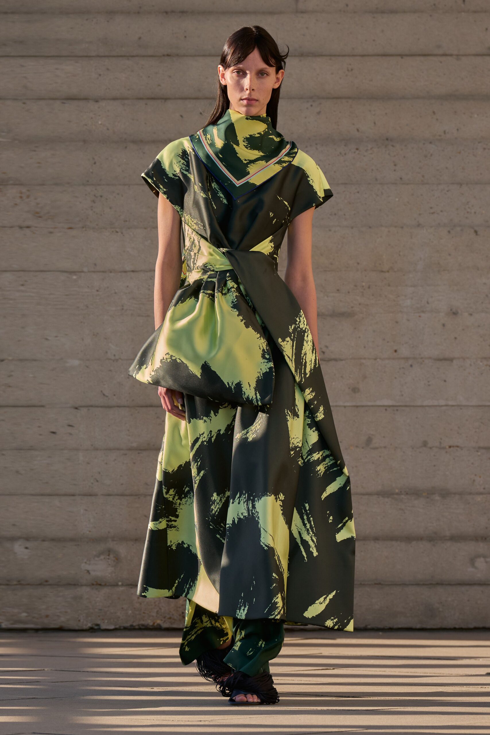 Chartreuse and pine satin scarf, striped border framing the point. Cap-sleeve satin dress in broad brushstroke print, one hip folded into a bow. Matching printed trousers peeking below the uneven hem. Black sandals shagged with fringe over the toes. Sun hitting courthouse steps after a rainless week.