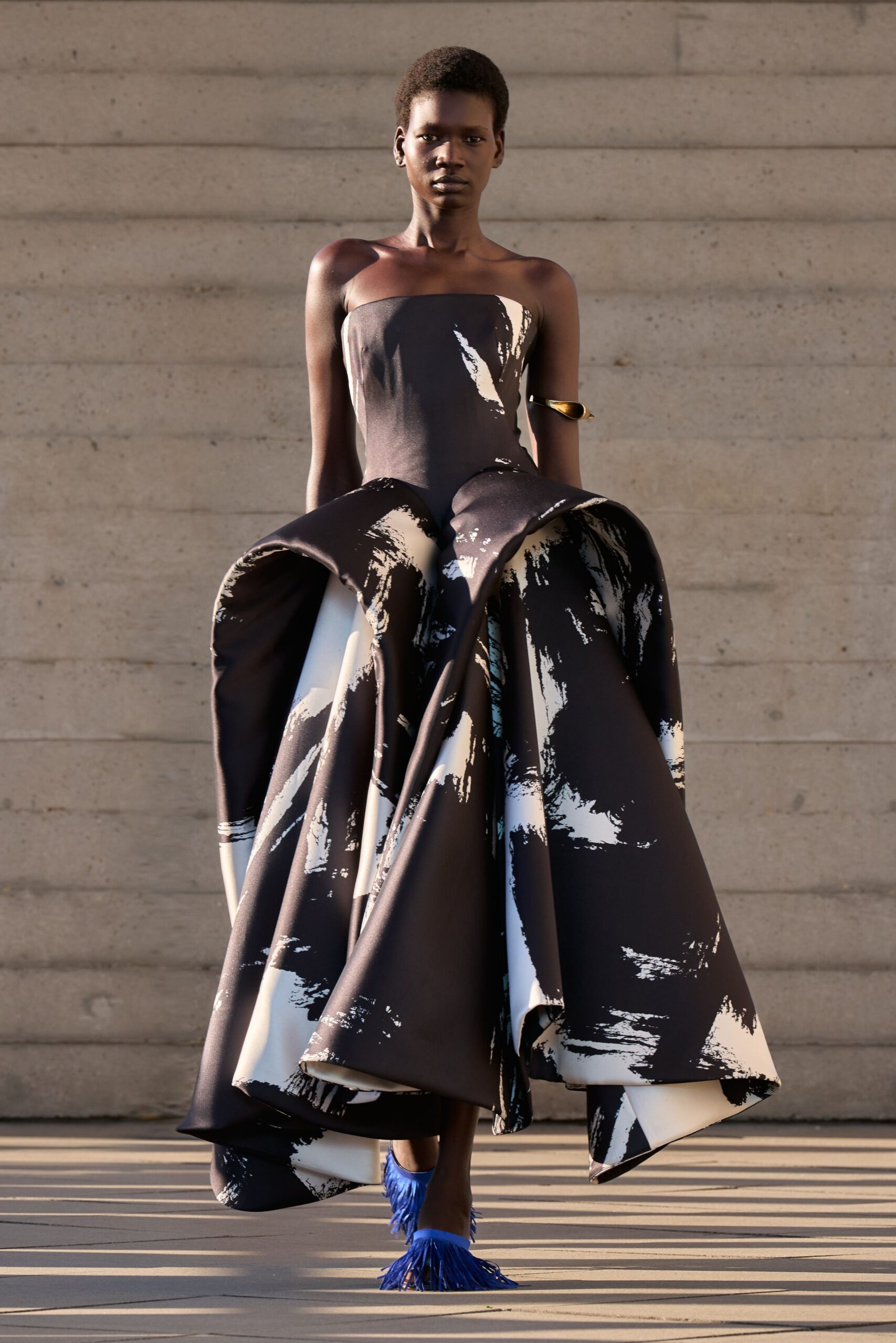 Strapless black satin gown splashed in white, hips built into vast folded panniers. Gold spiral cuff gripping the upper arm. Electric-blue mules with long raffia fringe skimming the pavement. Sun-struck museum plaza, everyone stops mid-conversation when she crosses.