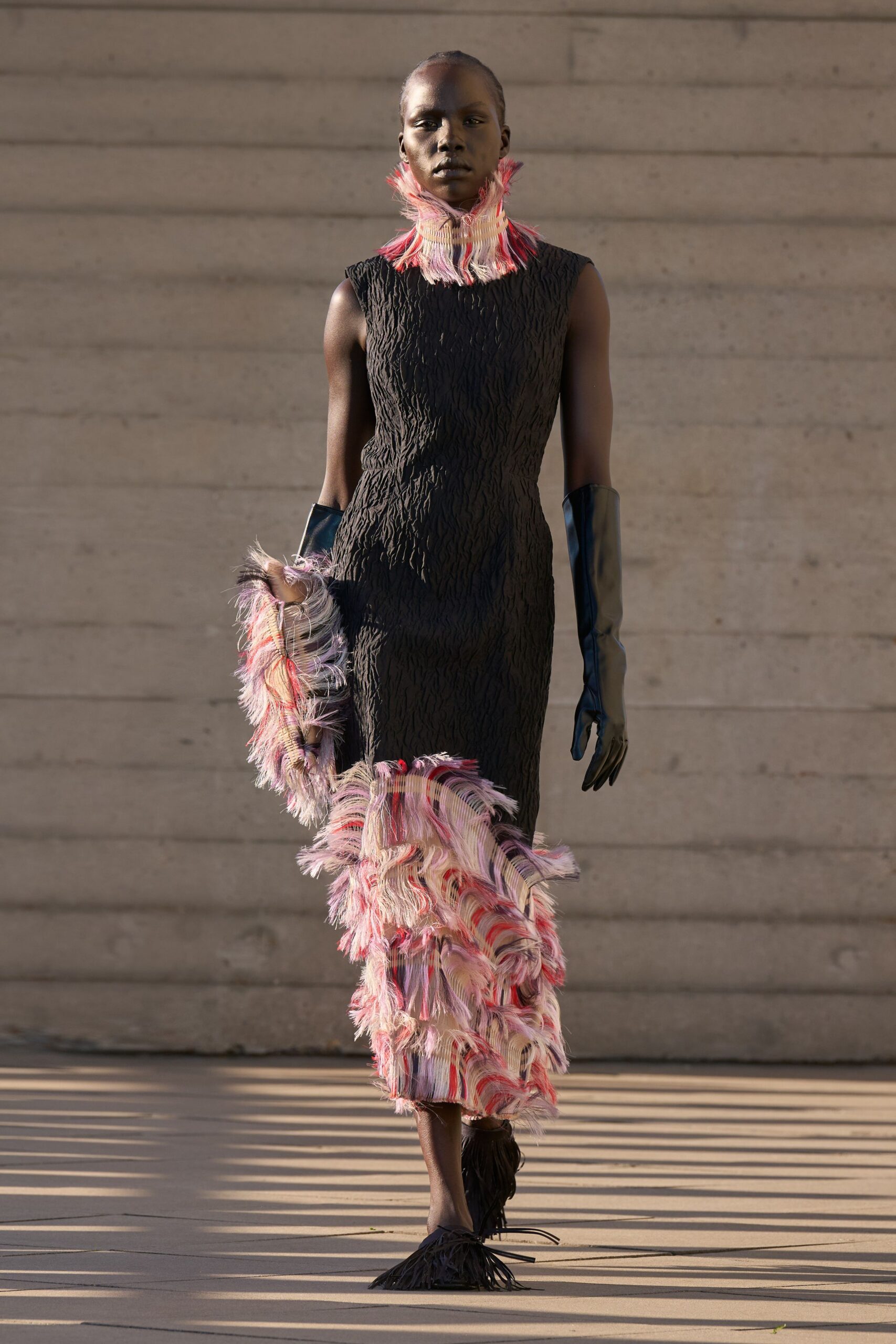 Tall fringed collar in pink, cream and red brushed threads. Black sleeveless column dress in puckered crinkle knit. Opera gloves in glossy black leather. Matching fringed scarf looped from hand to calf. Black flats with swinging fringe over the toes. Crossing a museum courtyard after the sun has dropped behind the concrete.