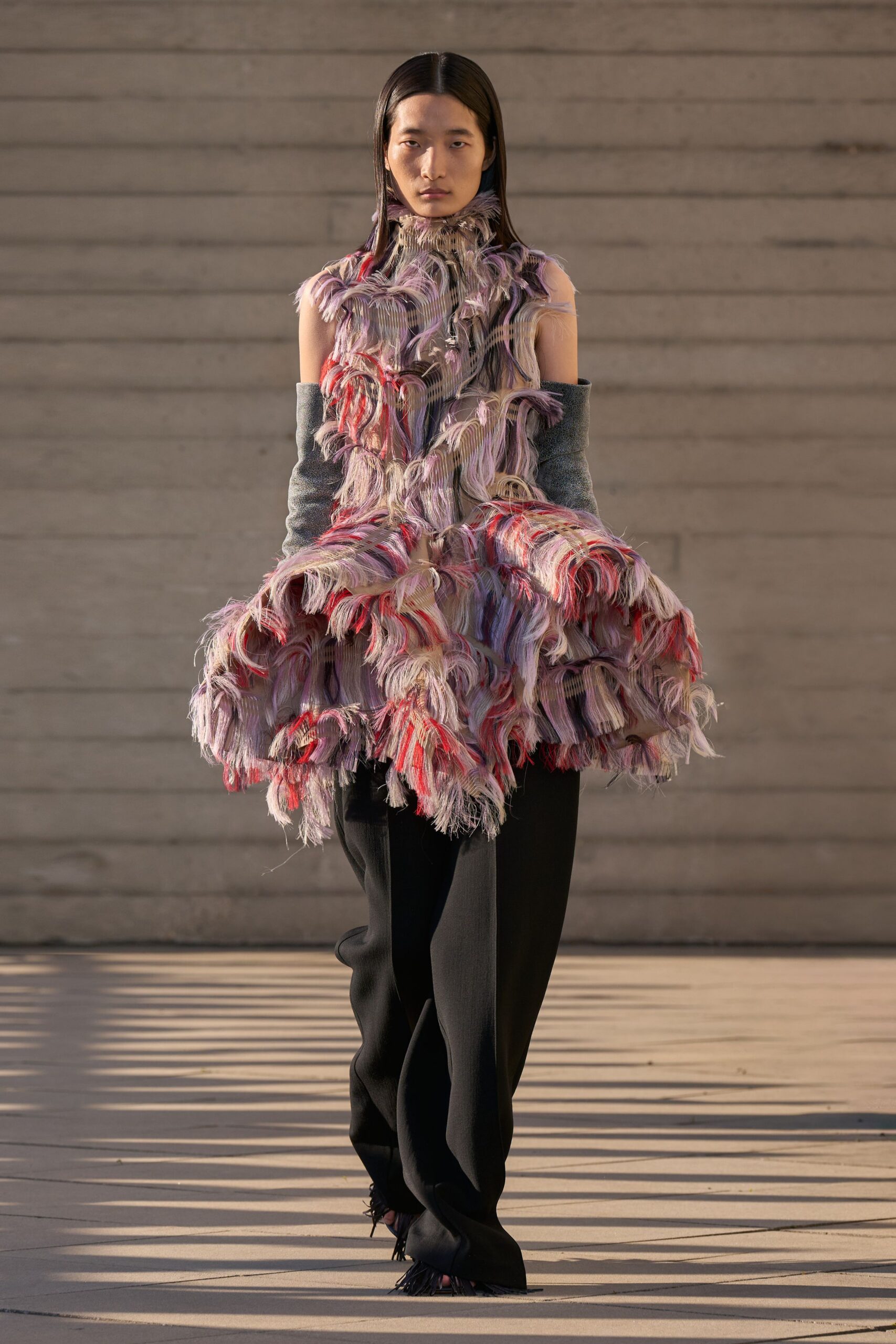 Sleeveless funnel-neck dress shagged with lilac, cream and scarlet looped fringe. Grey knit detached sleeves slouched below the bare shoulders. Black wide-leg trousers falling straight under the sculpted peplum. Black pointed shoes with fringe skimming the floor. Crossing a museum courtyard after wind lifts the fountain spray.