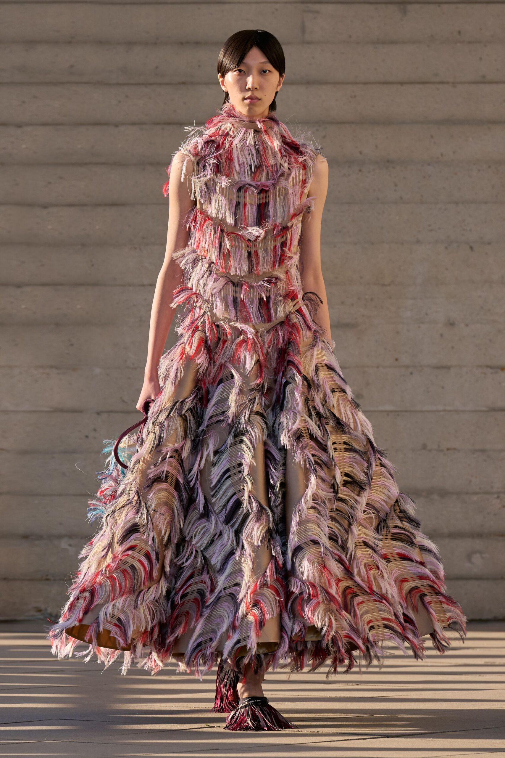 Sleeveless column opening into a full skirt, banded with long eyelash fringe in blush, plum and scarlet. Black sandals tufted with matching fringe over the toes. Sun on poured concrete, crossing a courtyard before the strings begin.