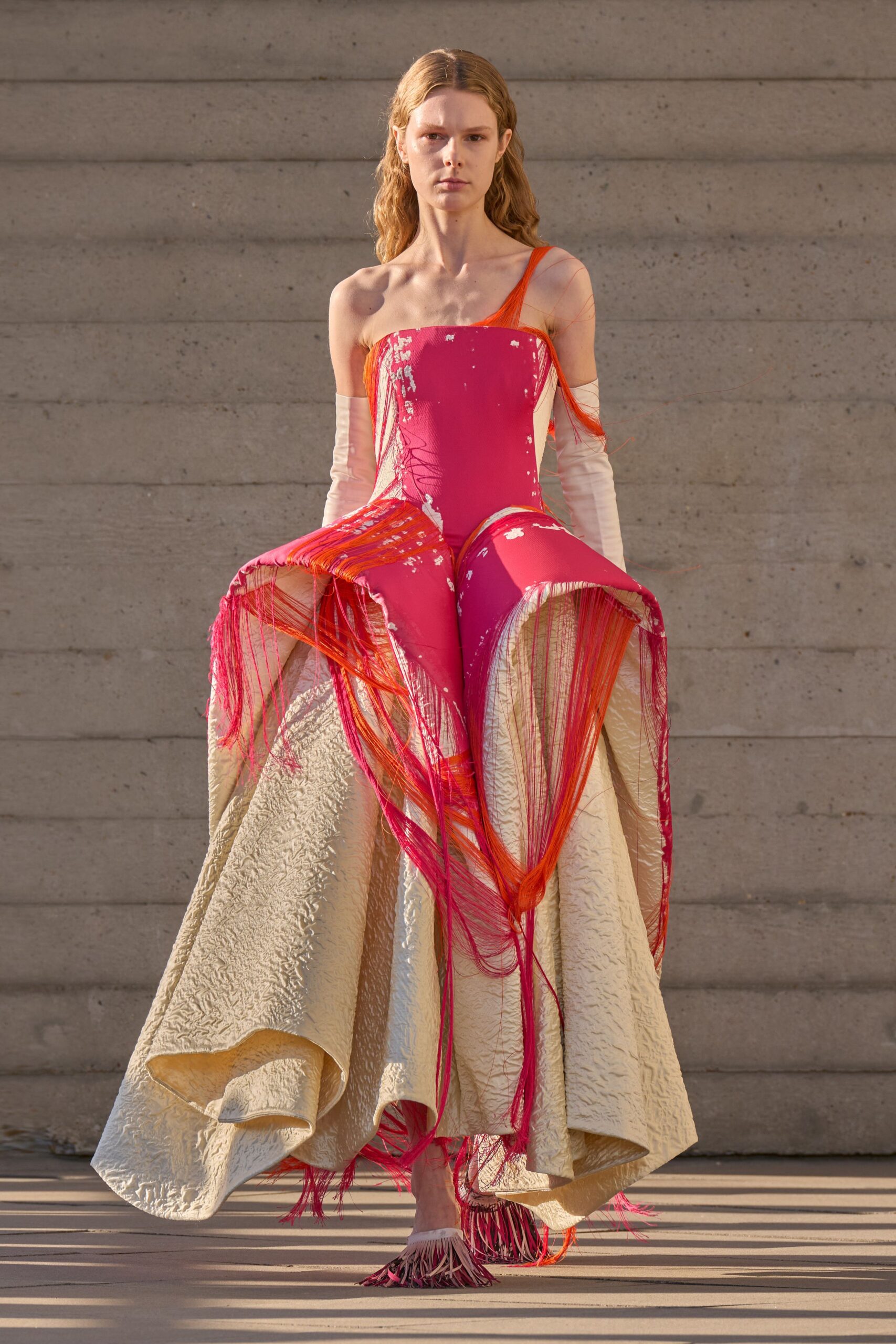 Hot-pink one-shoulder bodice exploding into a crinkled ivory bell skirt. Pale satin gloves slouched to the upper arm. Orange and magenta thread curtains knotted at the hips and hem. Pointed mules buried in swinging pink-and-cream fringe. Crossing a sculpture court just before the stone starts holding heat.