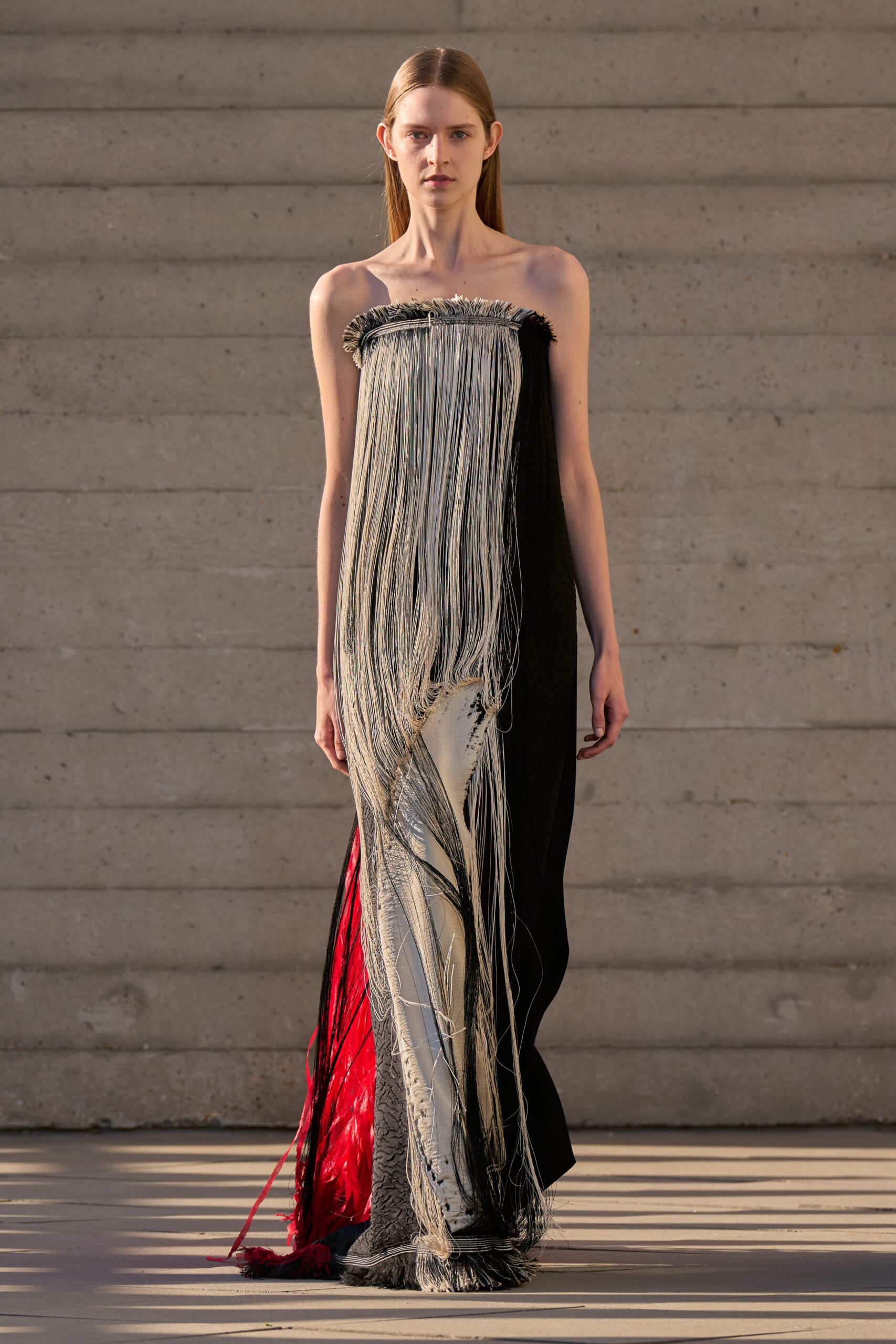Strapless column gown with a raw-fringed neckline and waterfall cord fringe. Black and ivory woven panel slit open, flashing a red underlayer. Sun hitting concrete steps after the ceremony has already emptied out.