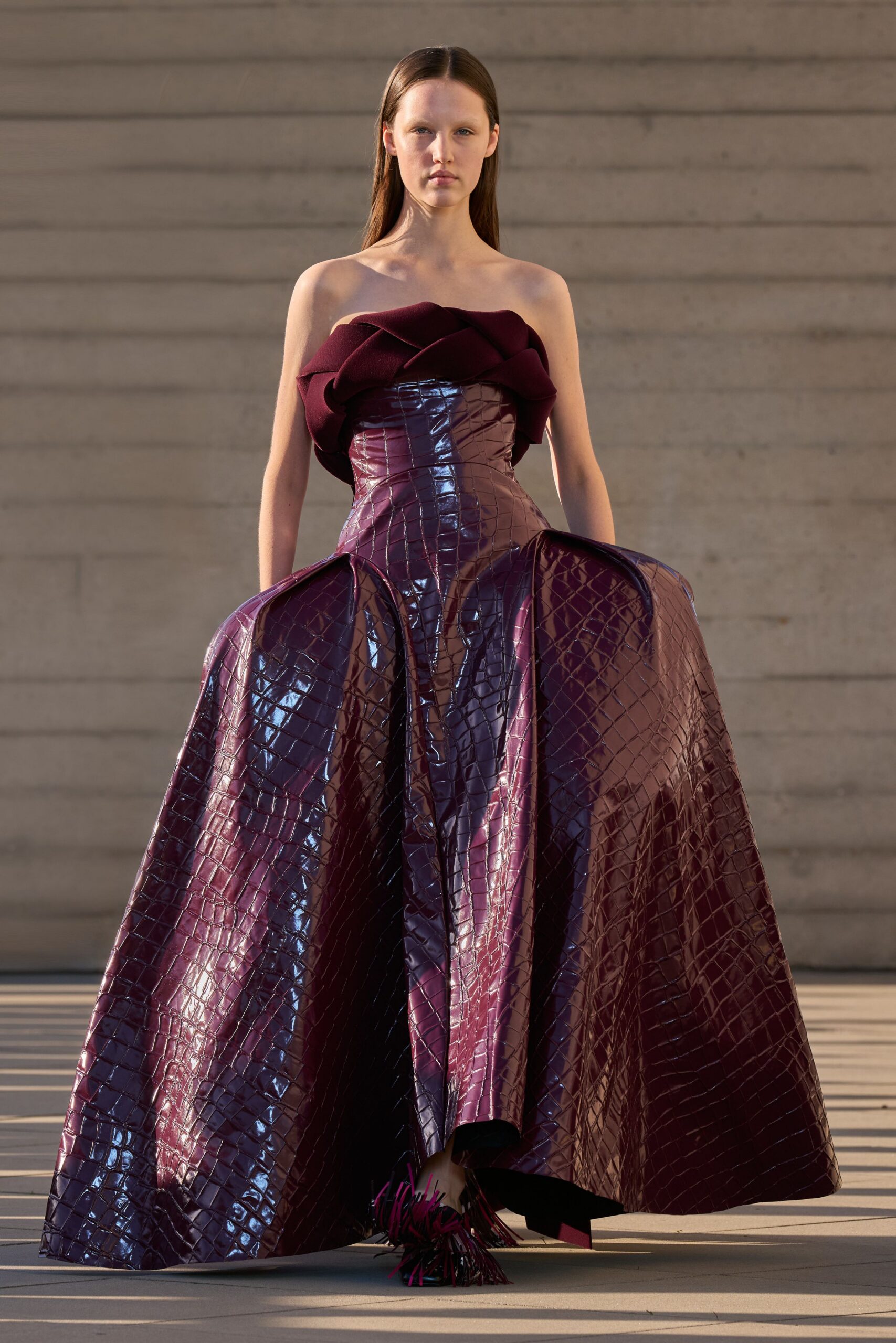 Strapless oxblood gown with a sculpted wool rosette neckline and mirror-shine crocodile embossing. Fringed wine heels peeking from the cavernous hem. Museum steps at sunset, after the speech has emptied the plaza.