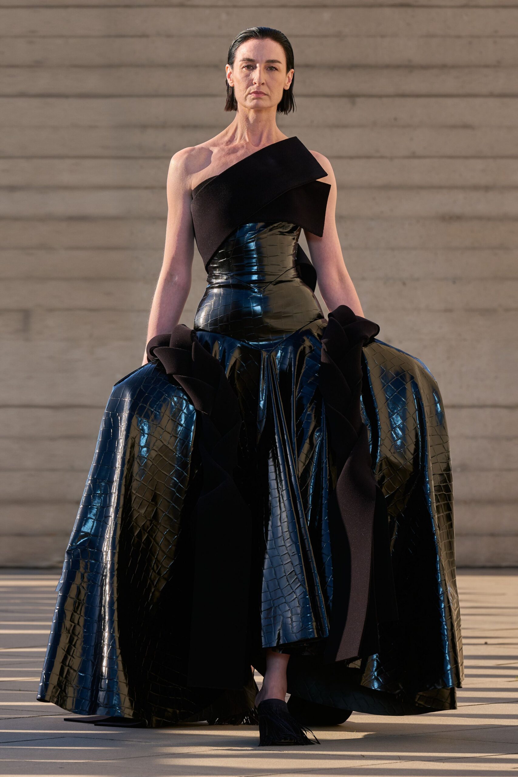 One-shoulder black gown, angular felt fold crossing a glossy crocodile-embossed torso. Vast patent crocodile skirt, split by matte ruffle spirals at each hip. Black flats with a fringe of thin cords skimming forward. Crossing a museum courtyard just before the donor dinner starts.