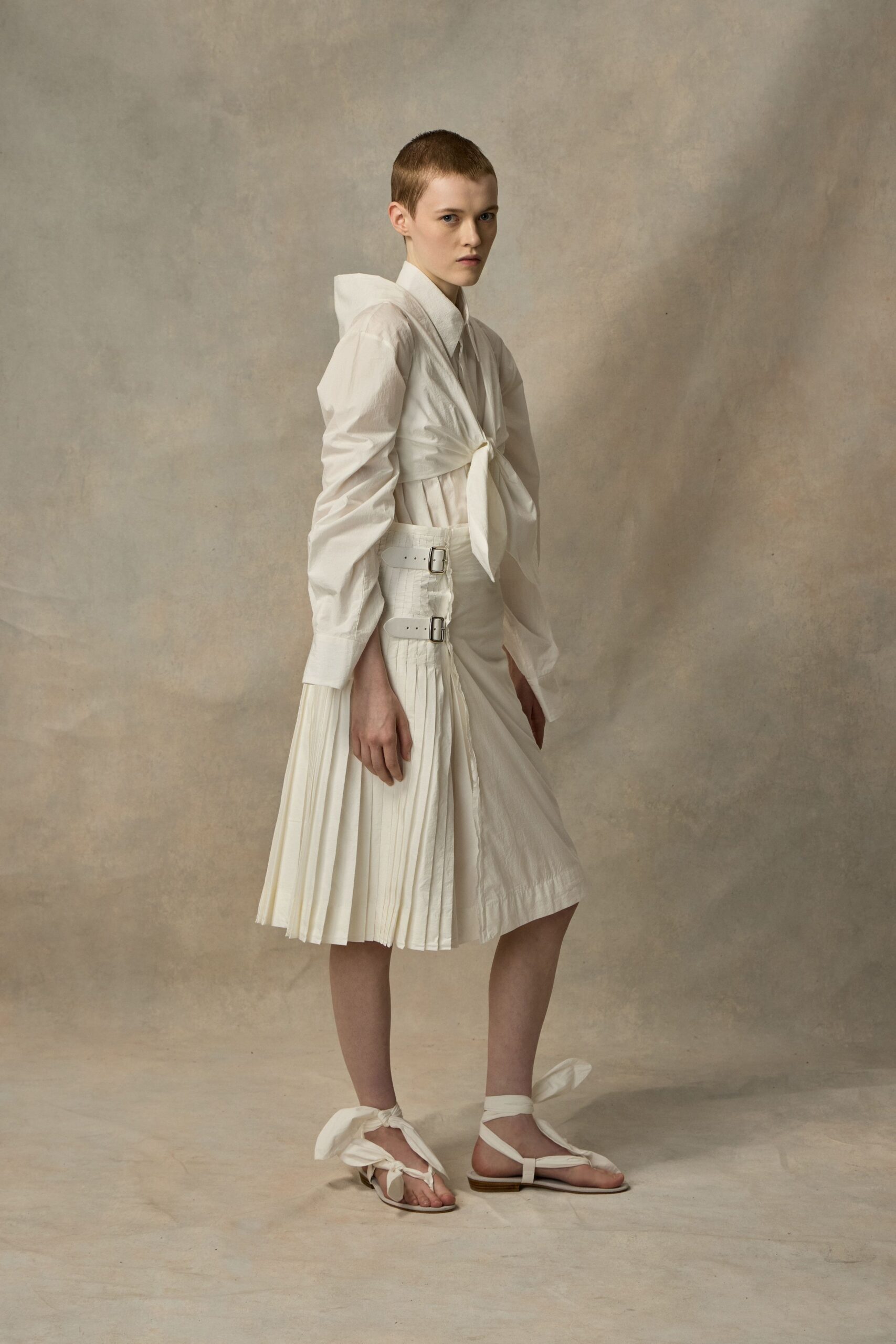 White cotton shirt with a high collar and elongated cuffs. Crisp white overlay tied at the bust, one shoulder sharply peaked. Midi skirt with knife pleats, side buckles and a split wrap front. Flat thong sandals laced with wide self-ties at the ankles. Crossing a cloister courtyard before the bells finish ringing.