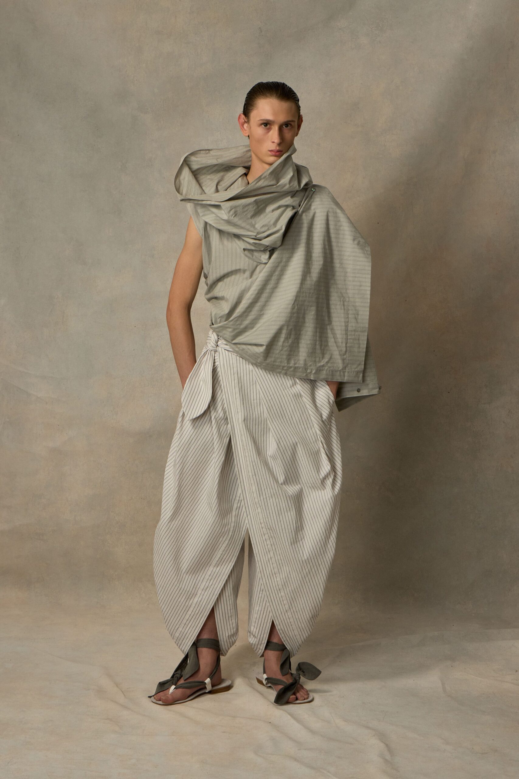 Pale grey shot cloth wrapped into a high funnel around the face. White pinstriped wrap trousers, one hip tied in a rounded flap knot. Flat thong sandals with charcoal ties climbing the ankles. Walking across sun-bleached flagstones before the museum doors open.