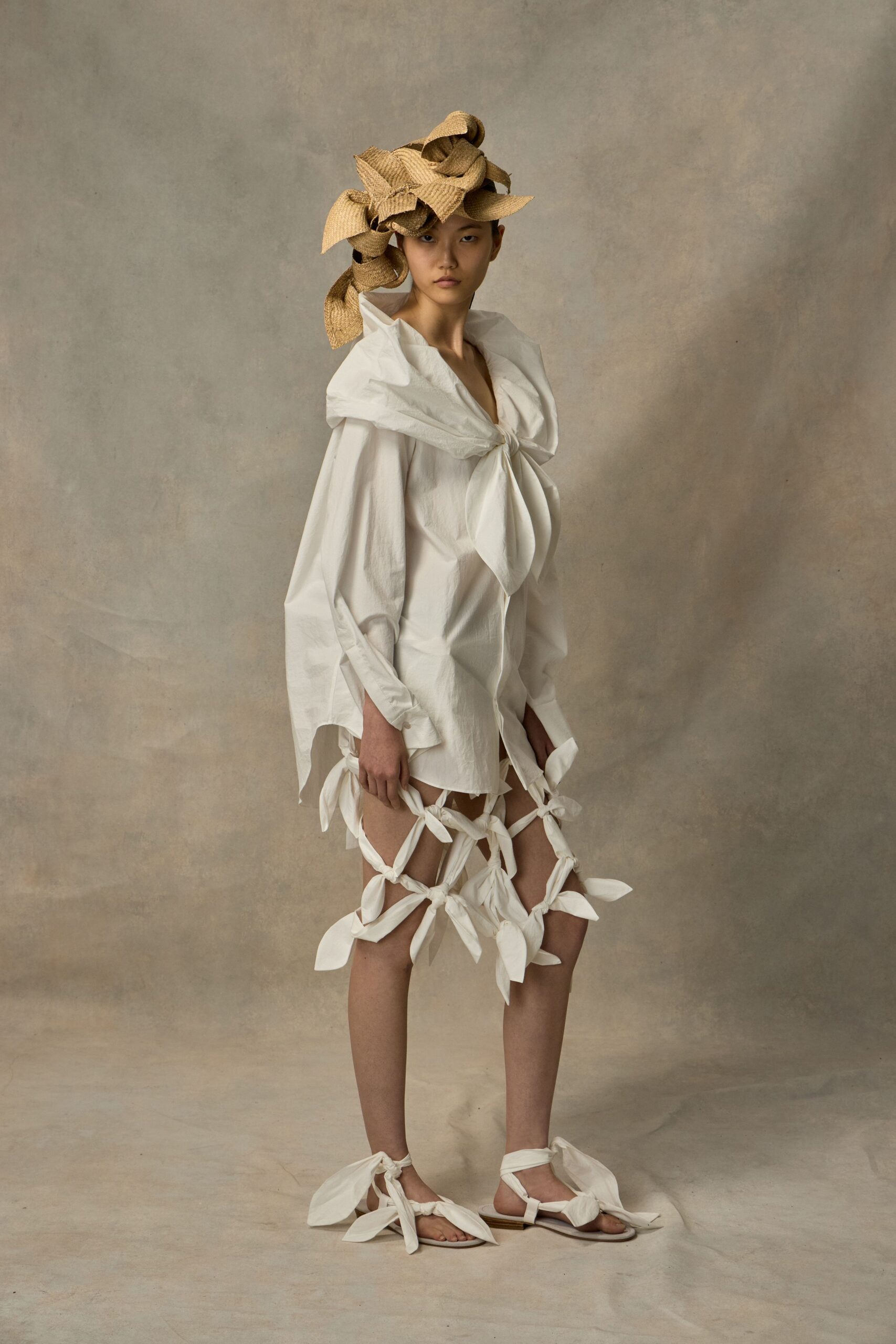 Crushed raffia hat spiraled into broad loops over one eye. White cotton mini with a giant shawl collar knotted at the chest. Matching fabric bands latticed up the thighs in petal-ended knots. Flat sandals tied with the same oversized white bows. For crossing a windy lawn after the ceremony, shoes sinking into the grass.