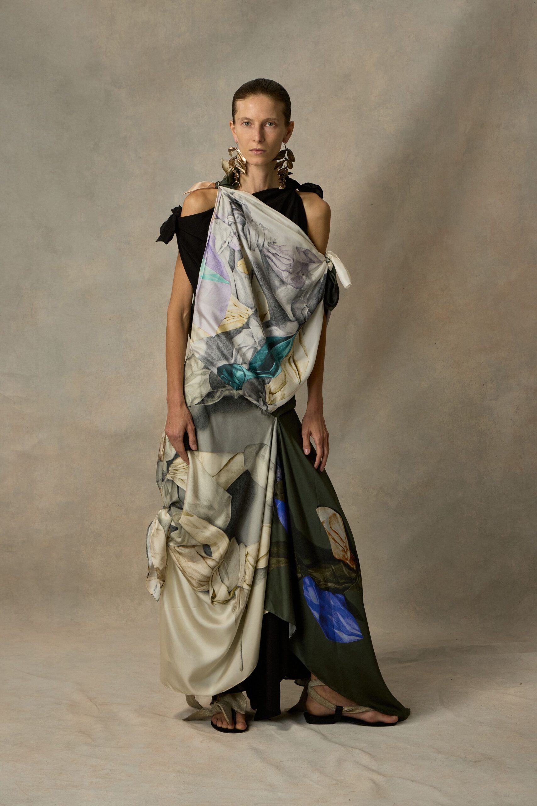 Long petal-cluster earrings in dull hammered metal. Black draped dress with tied shoulder tabs and a ruched side knot. Printed satin panel wrapped across the torso, falling in swollen folds. Flat thong sandals with pale suede straps. Museum atrium at closing, carrying your shoes in your hand by midnight.