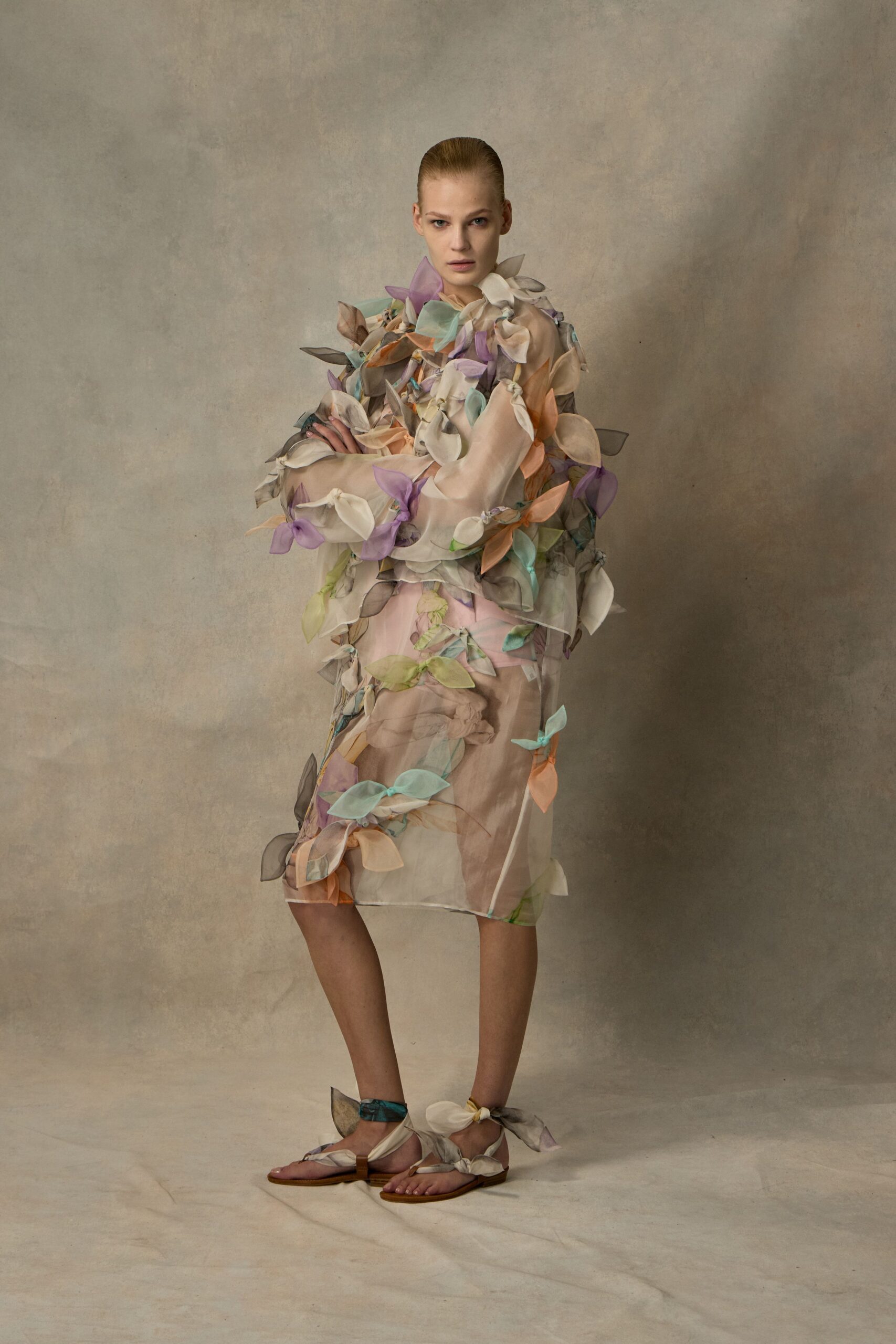 Sheer blush organza sheath covered in tied pastel petal appliques. Flat leather sandals wrapped with matching organza bows at the ankles. Greenhouse aisle after watering, petals stuck to your sleeves.