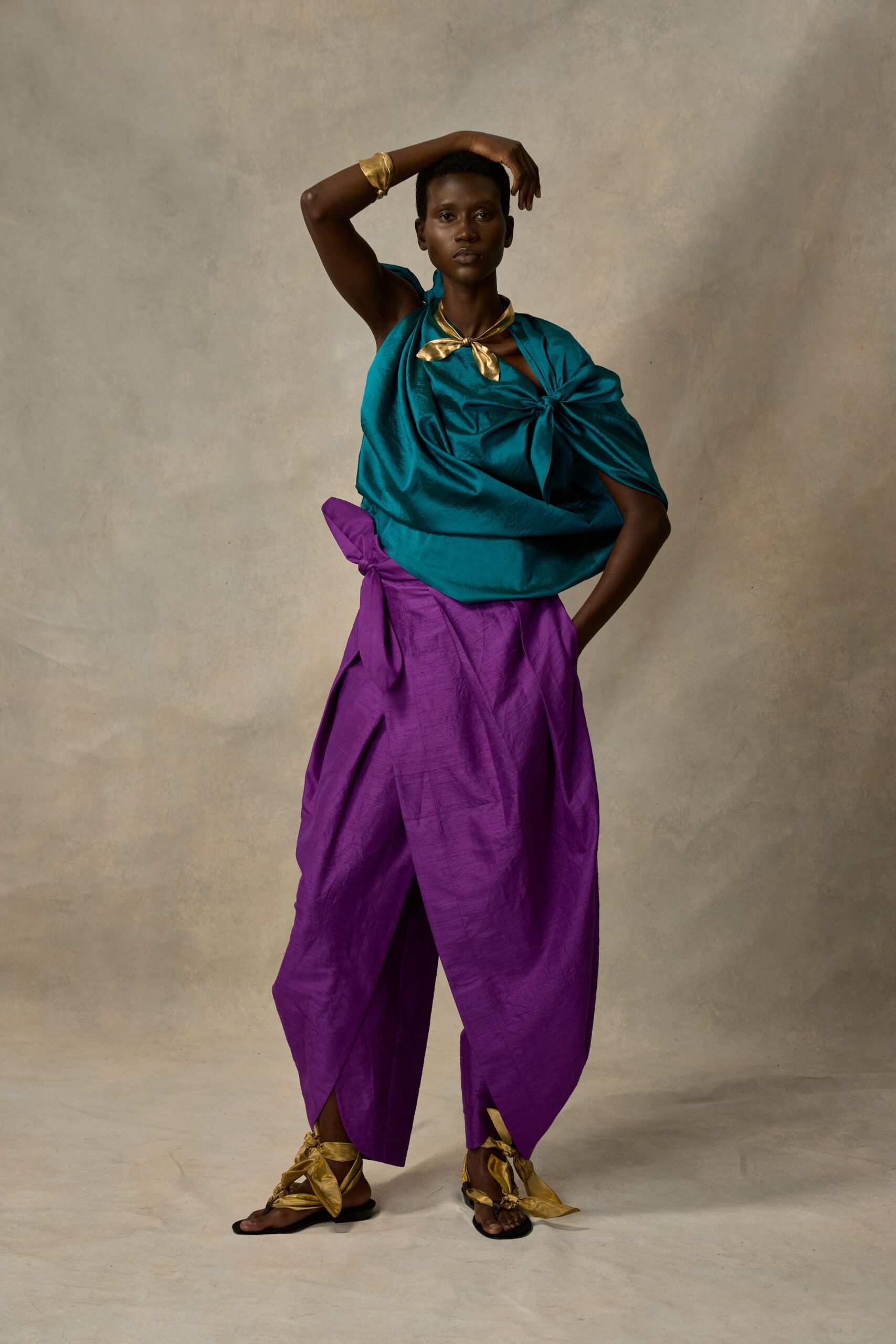 Gold satin scarf knotted close at the throat. Peacock satin top draped one-shoulder, gathered into a side knot. Hammered gold cuff. Amethyst silk trousers with wrapped tulip legs and a protruding hip bow. Gold thong sandals tied up in oversized satin bows. Walking into a courtyard wedding after the sun has dropped.