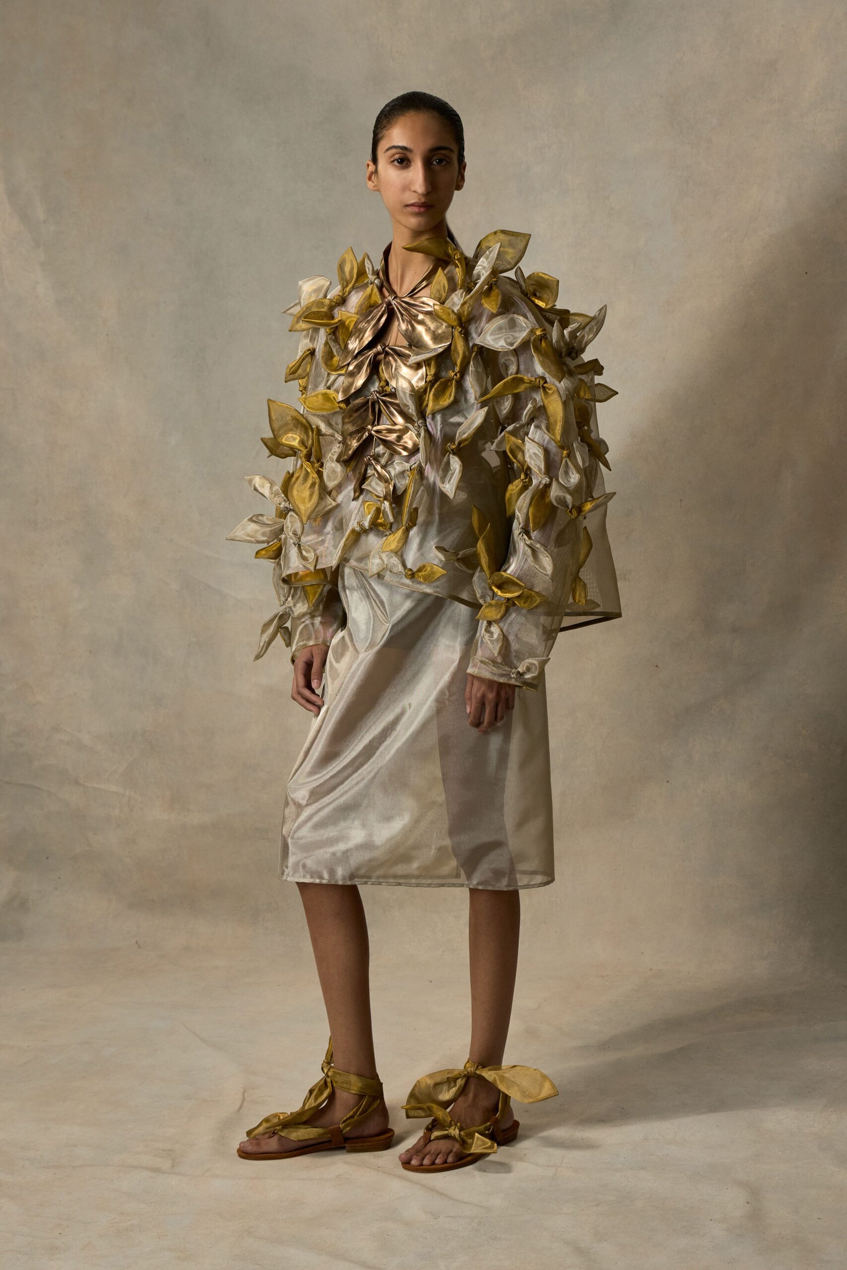Sheer organza jacket buried under padded gold and ivory leaf appliques. Pearly slip dress catching light in soft diagonal creases. Flat sandals tied with oversized metallic bows at the ankles. Greenhouse aisle after closing, watering cans lined up by the door.