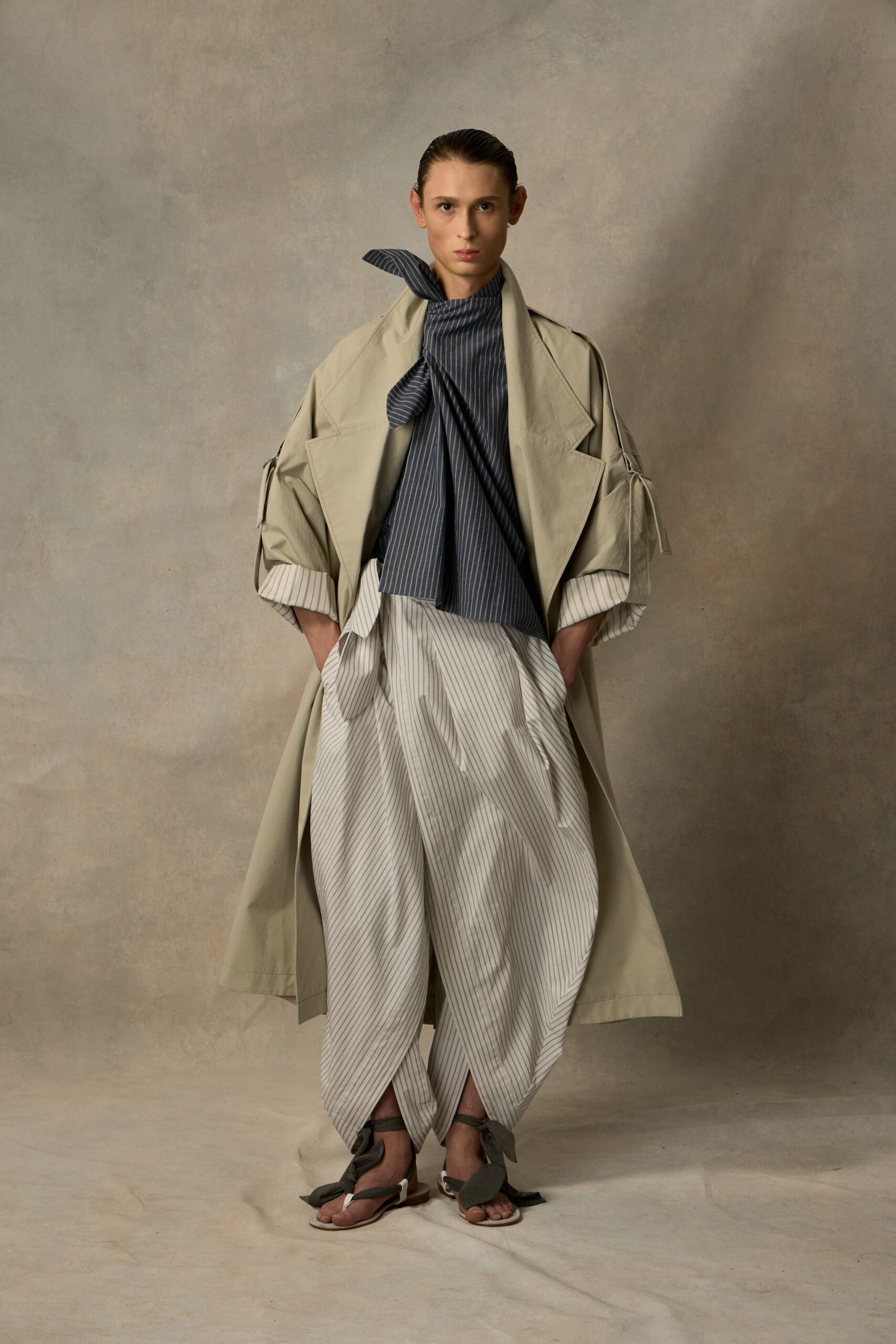 Stone cotton trench with cutout sleeves and striped cuff facings. Slate pinstripe scarf knotted high, one end kicked upright. Cream pinstripe wrap trousers crossing over the shins. Flat sandals tied with dark fabric bows at the ankles. Walking the tiled corridor between bathhouse rooms, robe forgotten somewhere behind you.