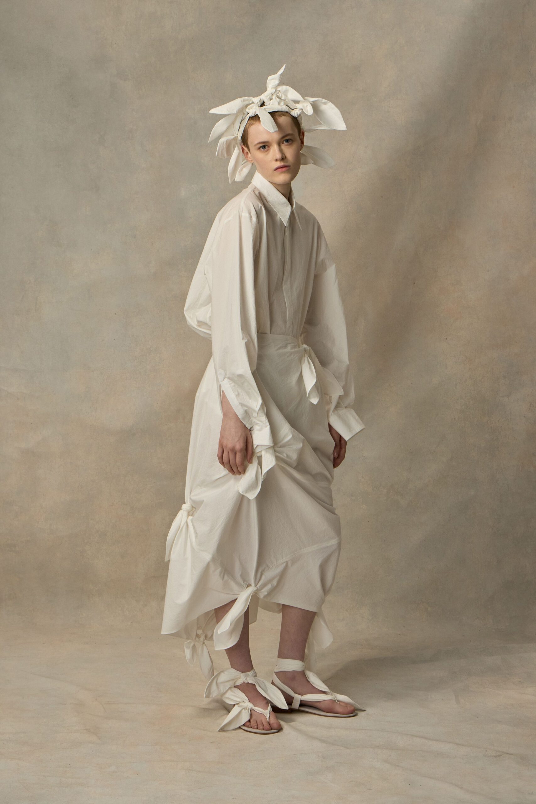 White cotton head wrap exploding into tied rabbit-ear loops. Crisp white shirtdress with dropped shoulders and a pointed collar. Matching cotton wrap skirt ruched into knots at hip and hem. Flat sandals laced with broad white ties around the ankles. Morning after the ceremony, crossing a tiled courtyard still carrying the bows.