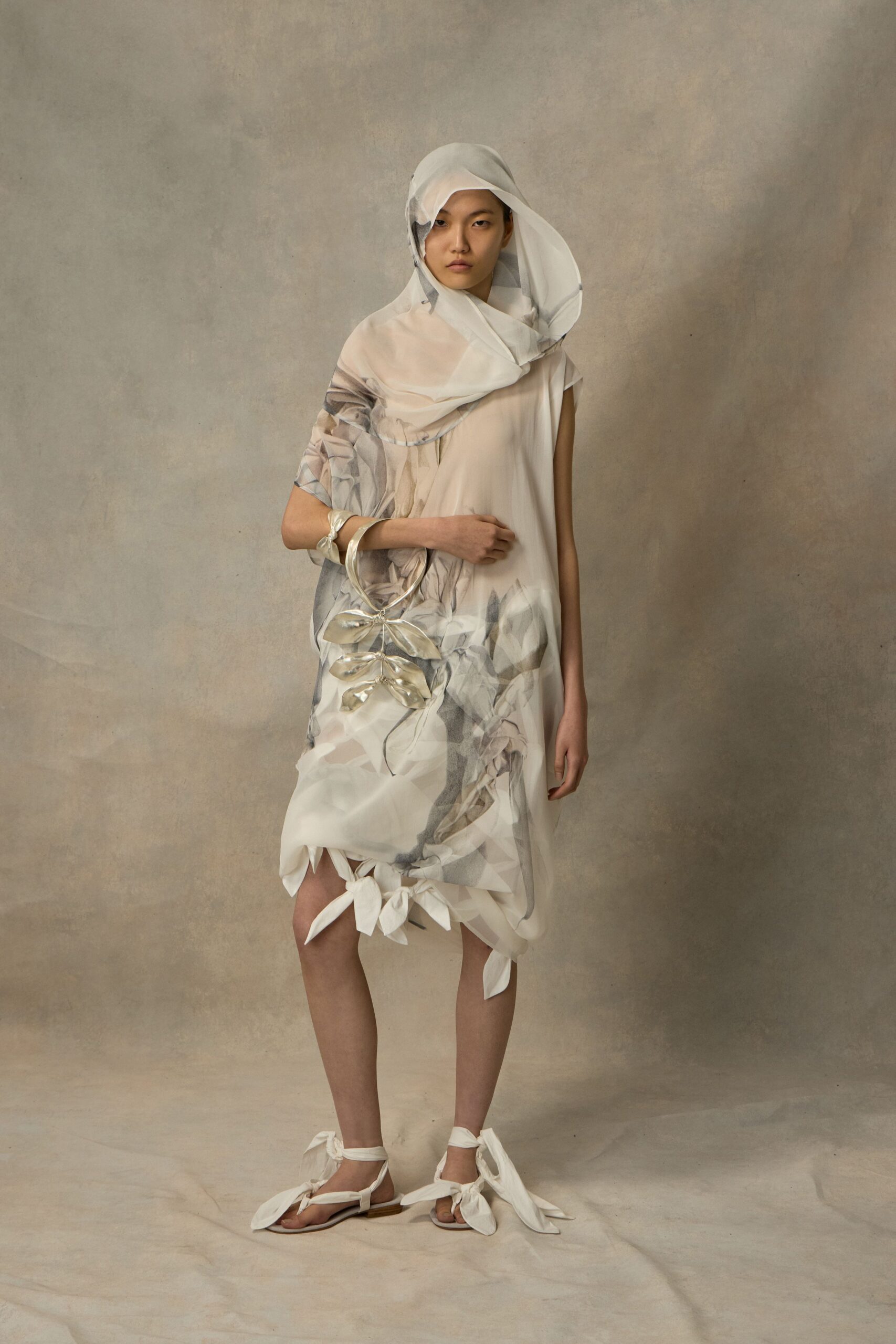 Sheer ivory hood scarf, wired edge folding into a soft oval. Cloudy floral chiffon shift, one cap sleeve and petal ties at the hem. Crinkled metallic cuff. Silver leaf-cluster ring handle bag. Flat white sandals knotted with oversized fabric leaves. Museum atrium at opening hour, florist buckets still sweating by the door.