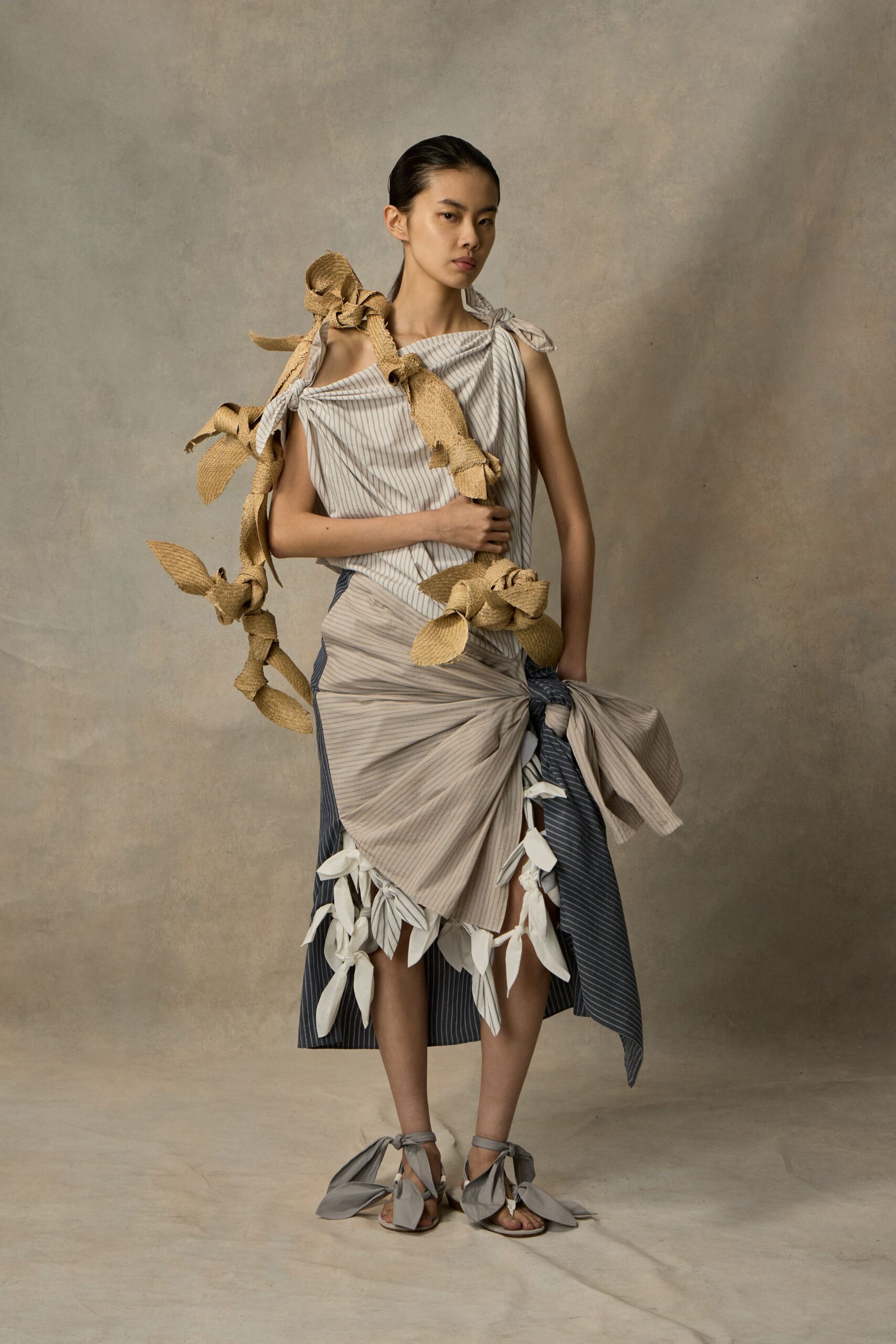 Asymmetric pinstriped wrap dress, tied at the shoulder and hip. White leaf-shaped tabs knotted all around the uneven hem. Looping burlap rope garland, frayed and tied in chunky bows. Flat sandals laced with oversized grey fabric ties at the ankles. Crossing a dry garden after the wedding chairs have been stacked.