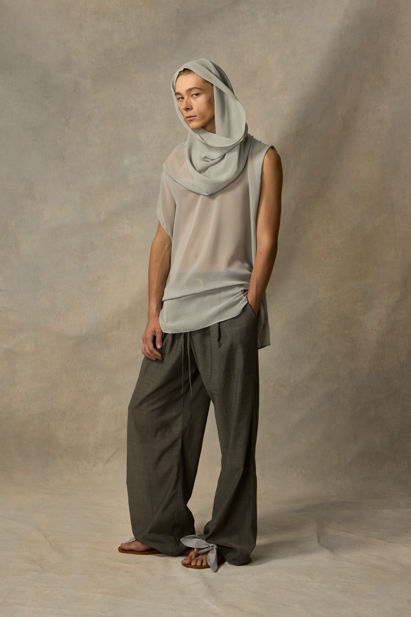 Pale dove drape wound into a soft hood and cowl. Sheer ice-grey tunic, one cap sleeve and one bare shoulder. Loose charcoal trousers with a dangling drawcord at the fly. Flat sandals tied over the toes with matching cloth. Crossing a hammam courtyard while the stone is still warm.