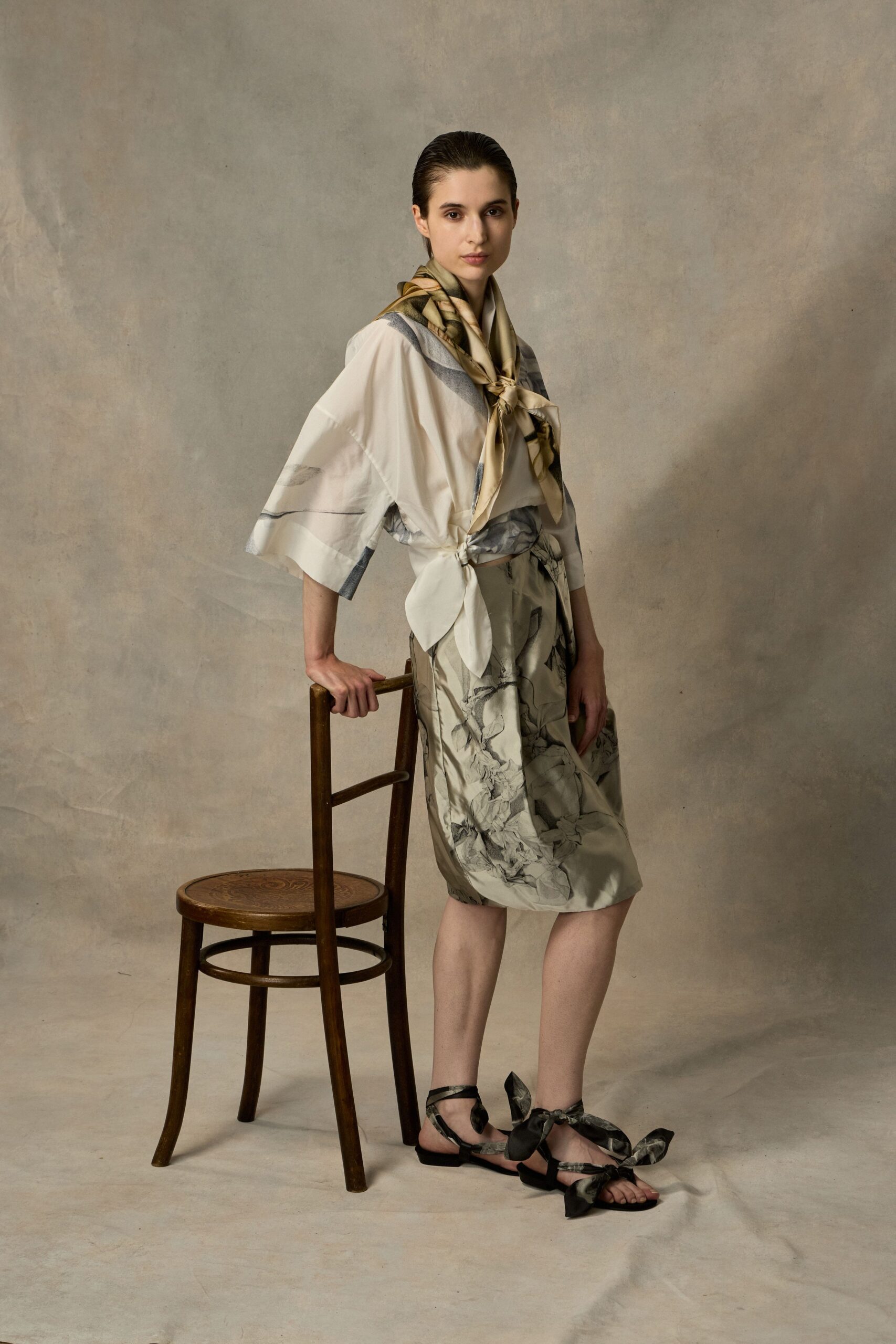Olive and cream silk scarf knotted low with a loose tail. Ivory wrap blouse in airy satin, wide sleeves cut square. Misty floral skirt with a tied side panel at the waist. Black flat sandals laced with oversized printed bows. In a quiet hotel suite before flowers arrive for the ceremony.