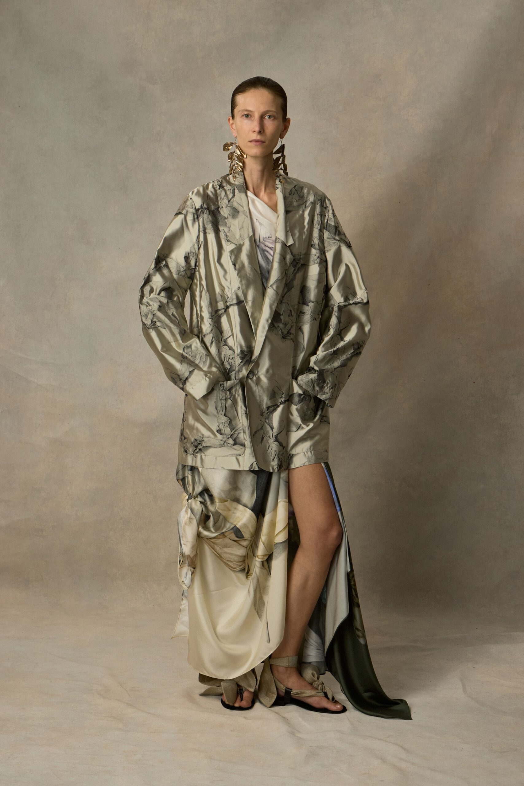 Long brass leaf clusters brushing the neck. Pearl-grey satin blazer printed with charcoal branch tracings. Ivory draped satin dress, one side knotted into a heavy swag. Flat sandals with champagne ties winding the ankles. Museum courtyard at dusk, carrying your shoes by dessert.