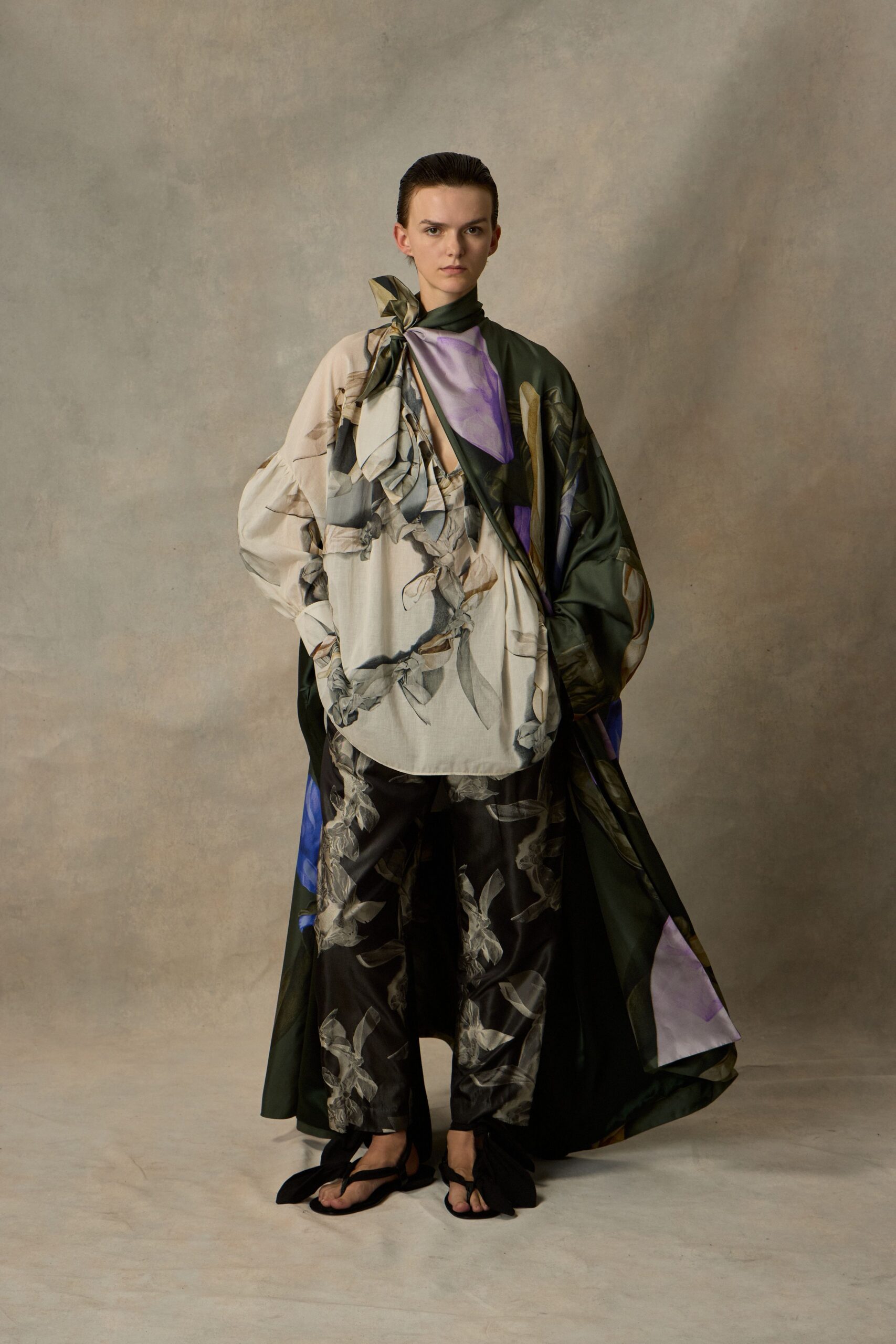 Dark olive satin scarf coat knotted high at one shoulder. Ivory blouse with balloon sleeves and washed iris print. Black satin cropped trousers scattered with ghostly petal motifs. Flat black sandals with long ties pooling around the ankles. Walking across a museum courtyard after rain, brochure still folded in your hand.