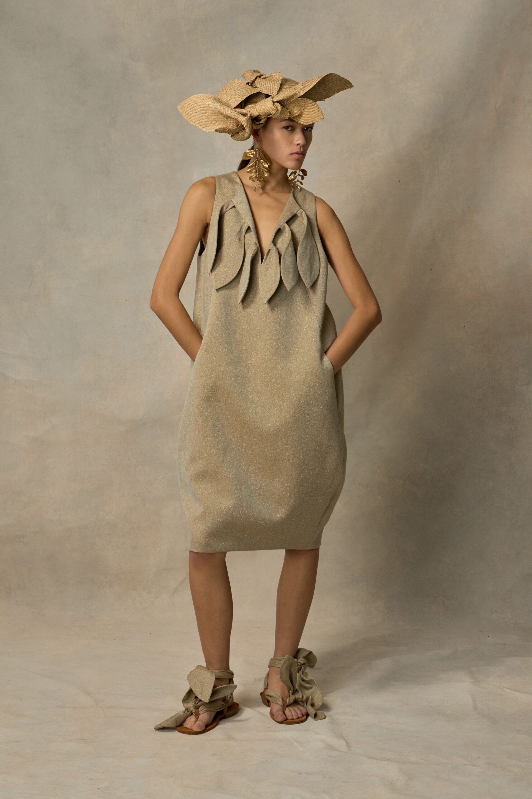 Oversized raffia bow hat, loops jutting out in uneven planes. Gold leaf-drop earrings brushing the jaw. Oatmeal shift dress with padded leaf appliques around a deep V. Flat sandals wrapped in giant tied fabric bows at the ankles. For a garden lunch where the tablecloth keeps lifting in the wind.