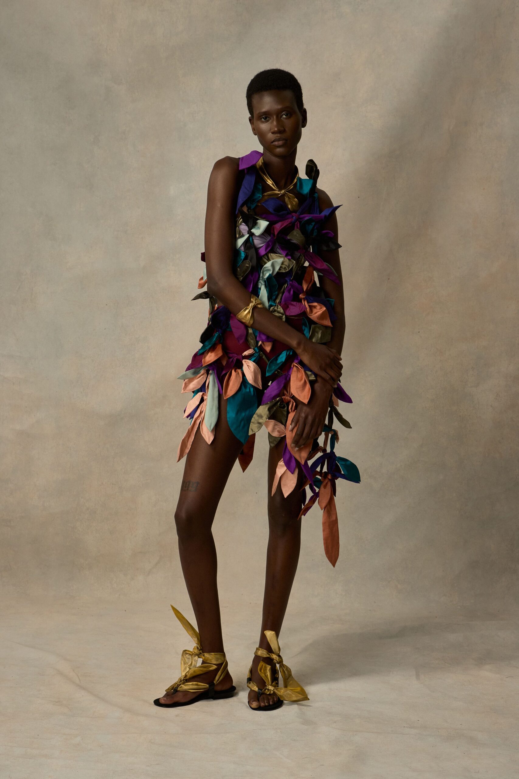 Sleeveless shift buried under satin leaf appliques in teal, plum, blush and olive. Metallic gold scarf knotted close at the throat. Crushed gold cuff twisted soft around the wrist. Flat black sandals laced with oversized gold satin leaves at the ankles. Walking out of a winter-garden party with petals stuck to everything.